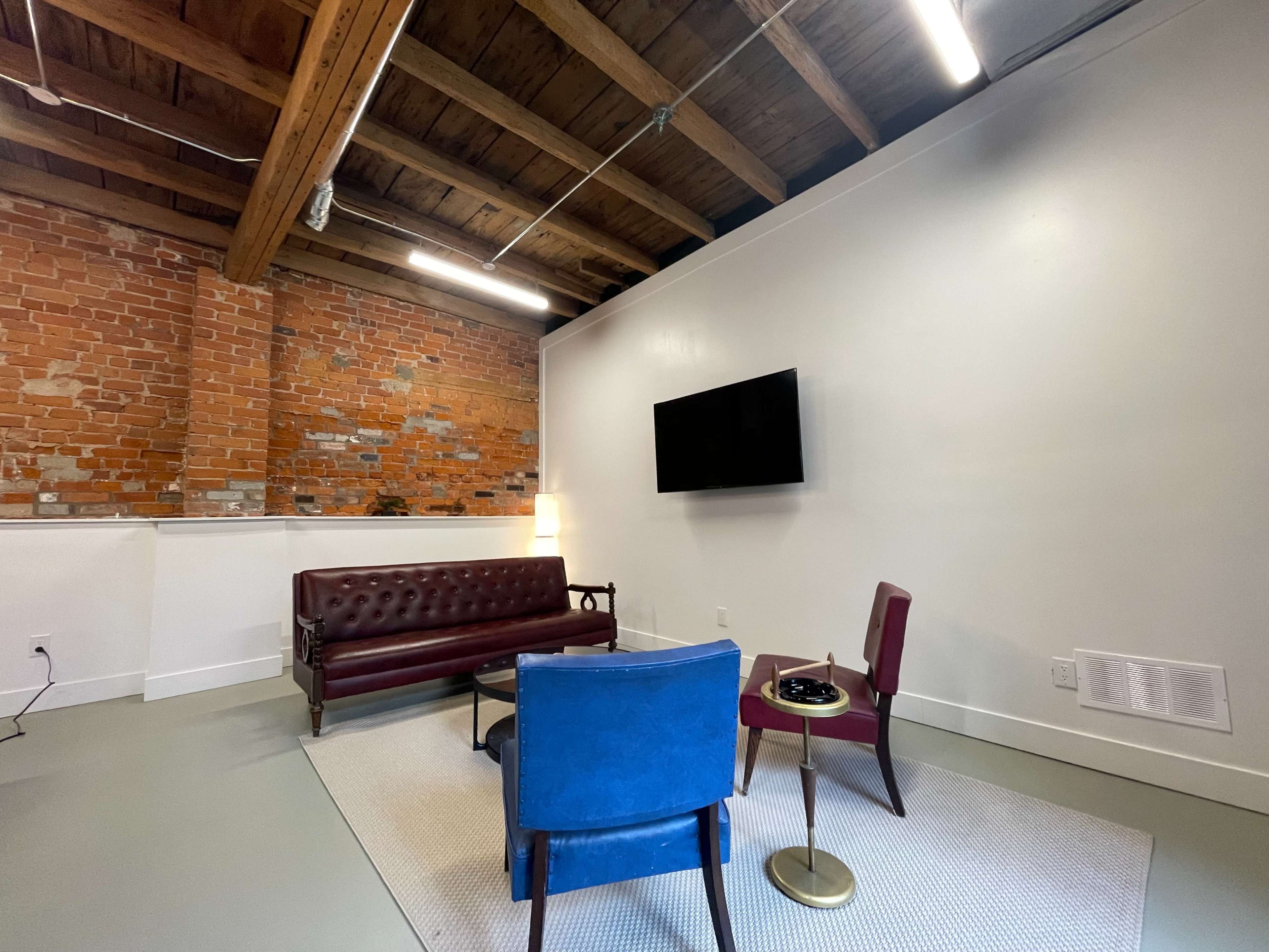 A minimalist lounge area featuring a brown leather sofa, two chairs, a small table, and a large wall-mounted TV against a backdrop of exposed brick and wooden beams.