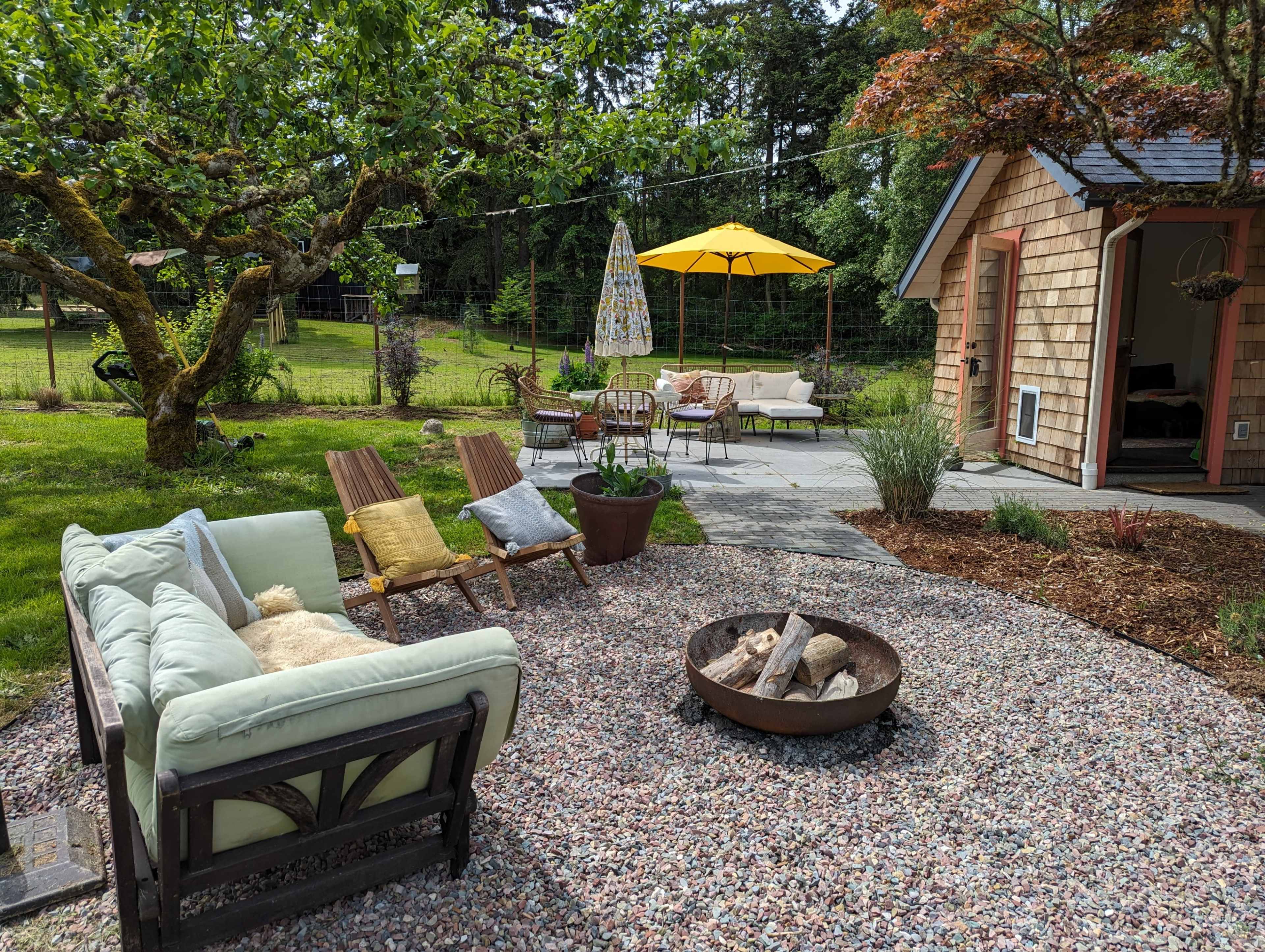 The scene features a cozy outdoor seating area with a fire pit, surrounded by greenery and a small, wooden shed.