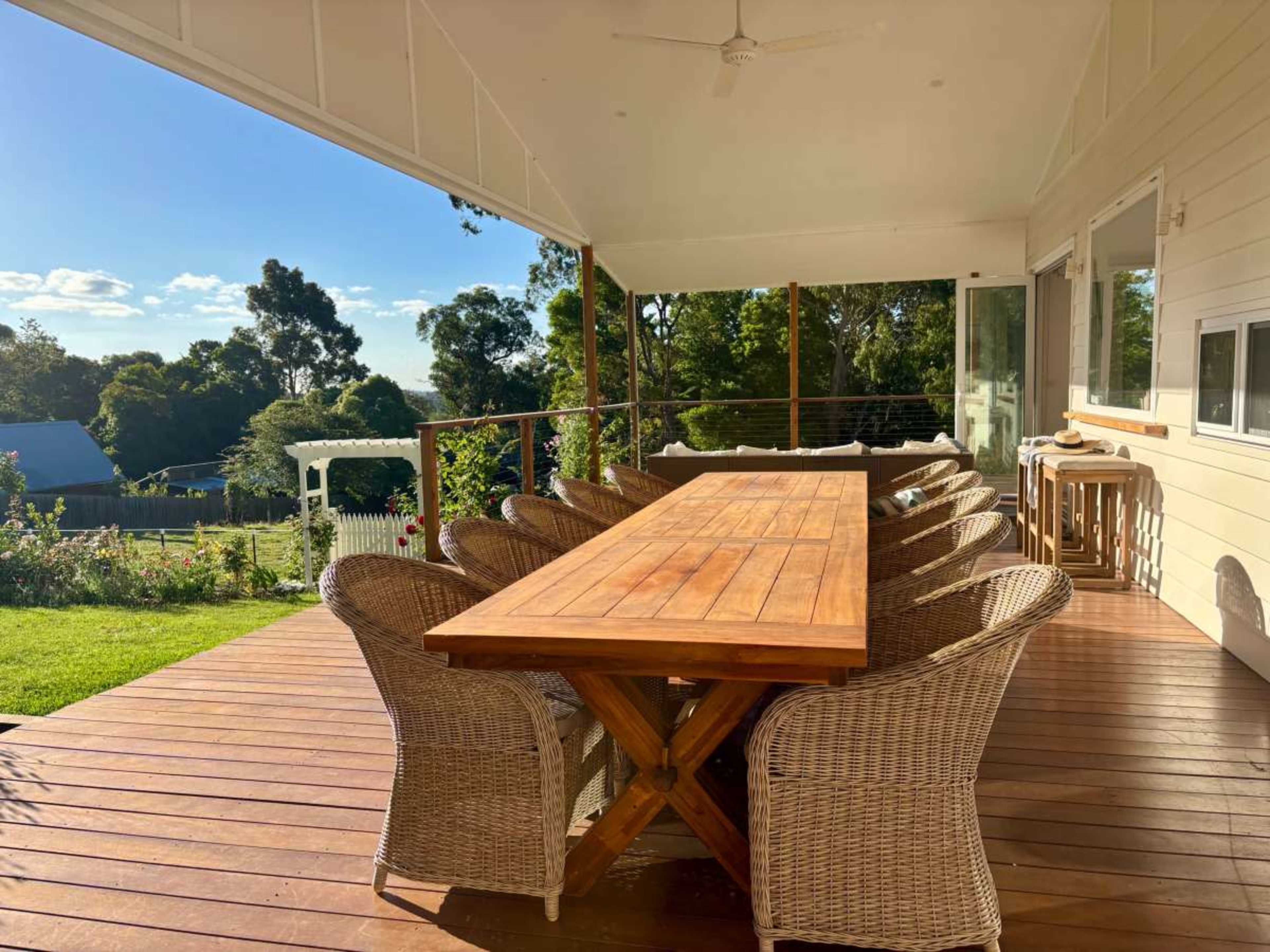 A large wooden dining table is set on a deck surrounded by wicker chairs, overlooking a green landscape under a clear blue sky.