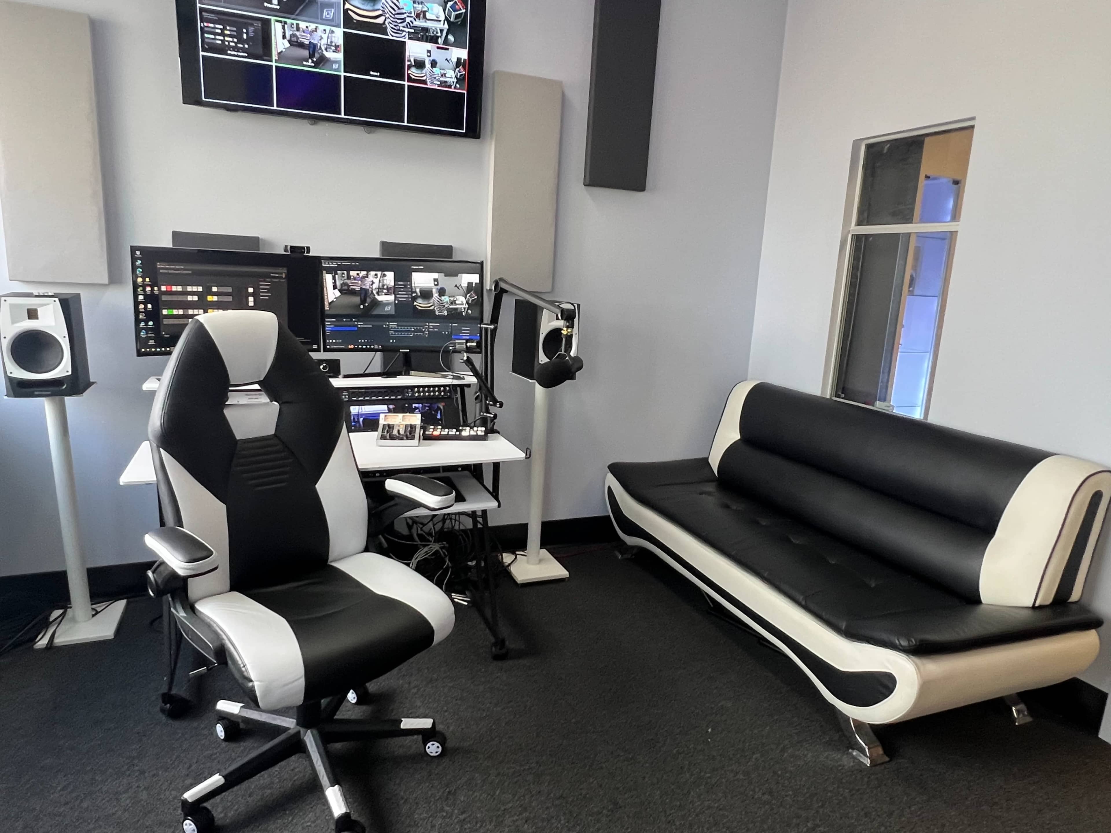 A sleek modern workspace featuring a desk with dual monitors and audio equipment, a black and white ergonomic office chair, and a black sofa against the wall.