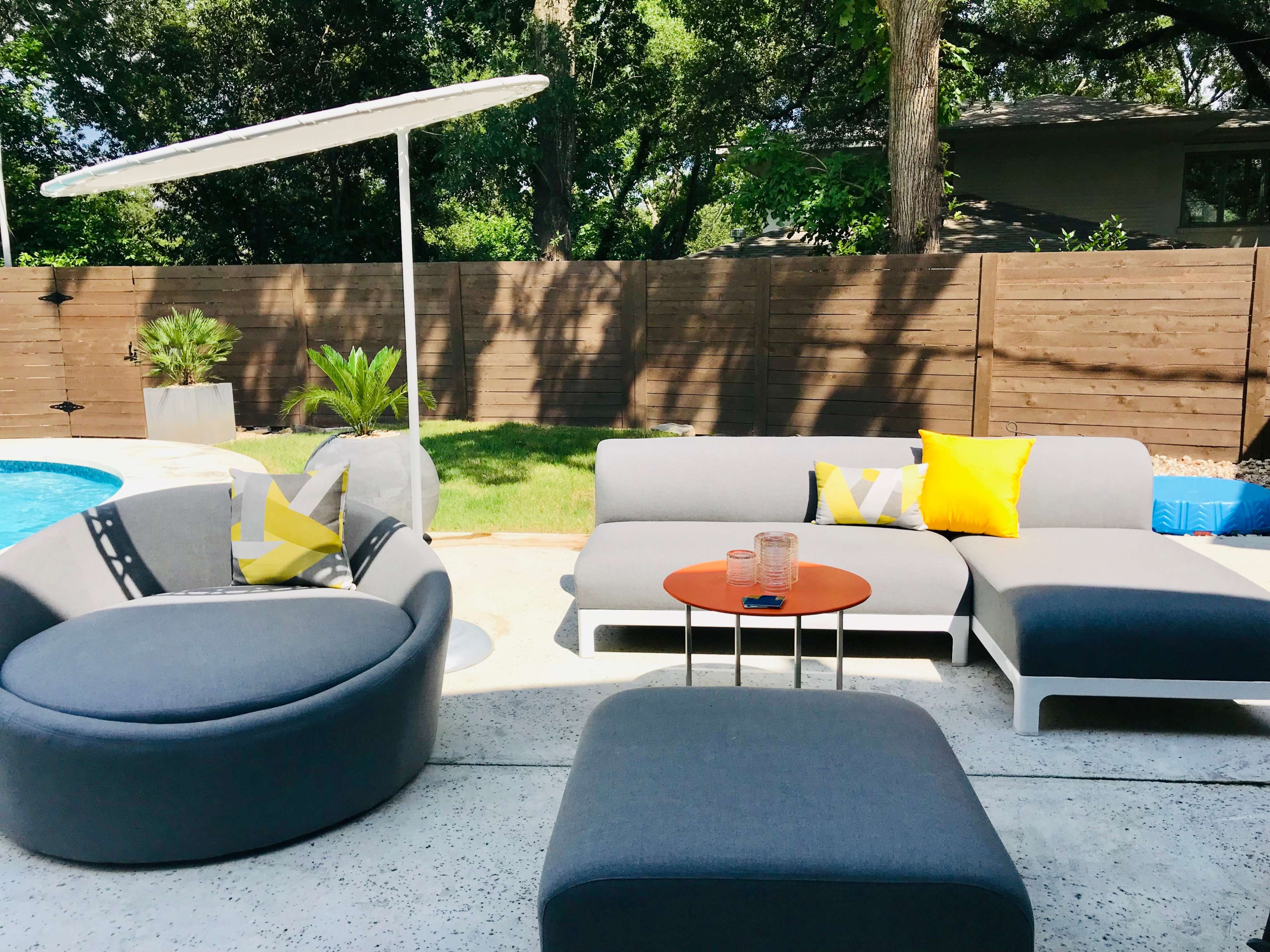 The scene showcases a modern outdoor patio with gray furniture, a small orange table, and a pool surrounded by lush greenery.