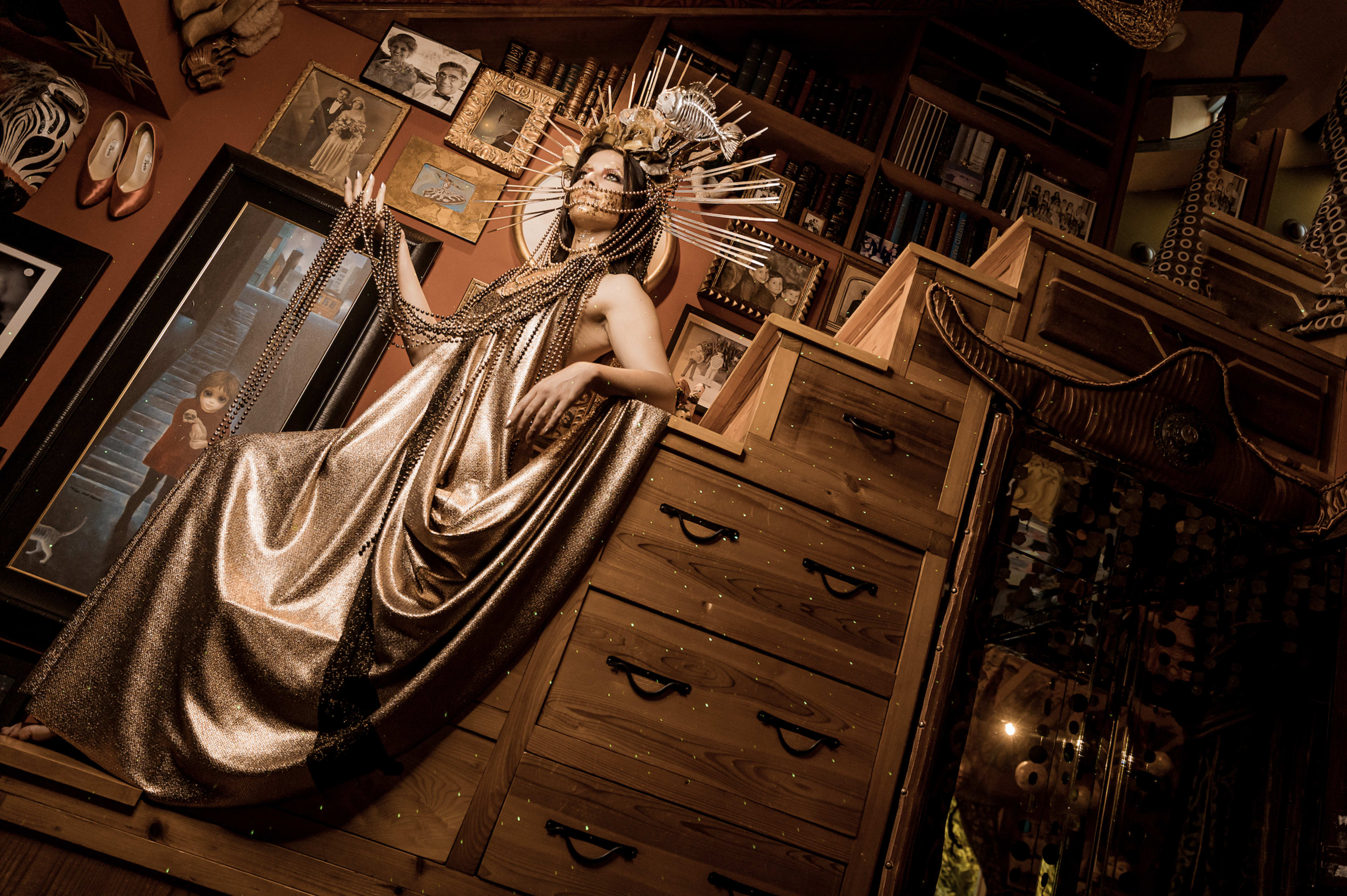 A figure dressed in a shimmering gold gown sits atop a wooden dresser surrounded by eclectic decor and books.