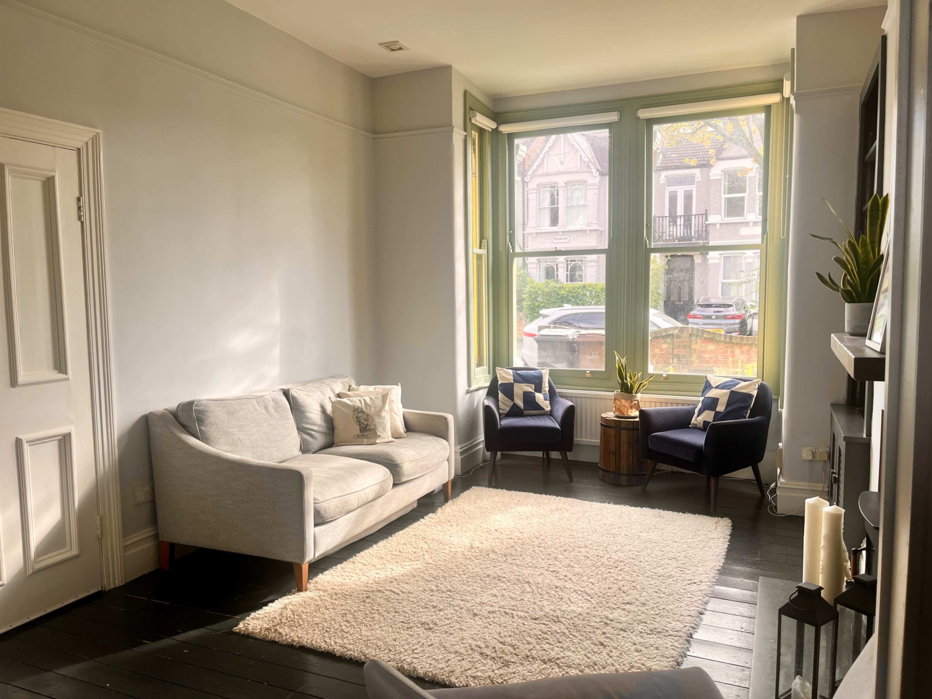A bright living room features a light gray sofa, two dark blue armchairs, and large windows overlooking a neighborhood street.