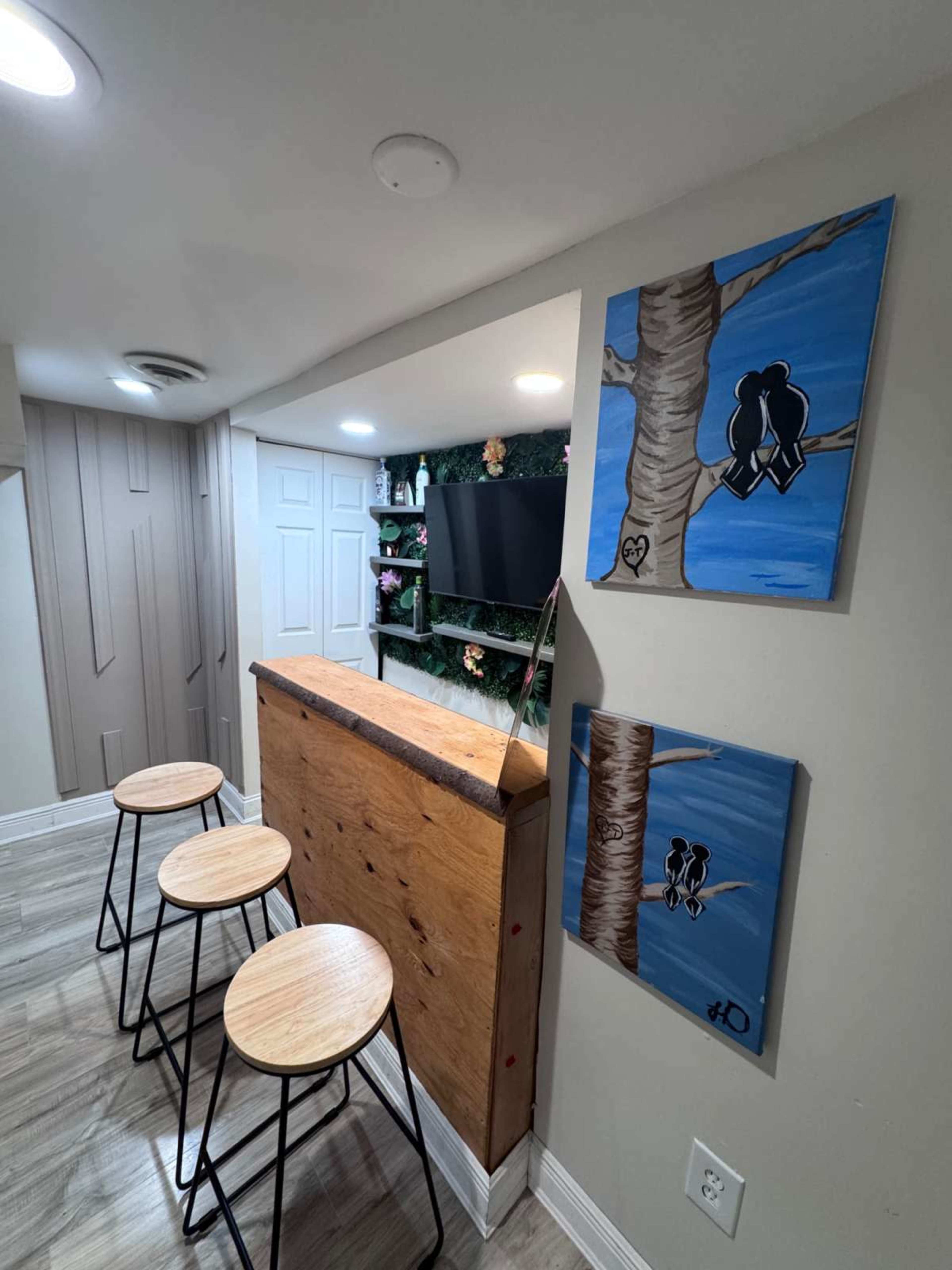The image shows a cozy basement bar area featuring a wooden counter, three stools, a wall-mounted television, and two framed paintings of birds on a tree.