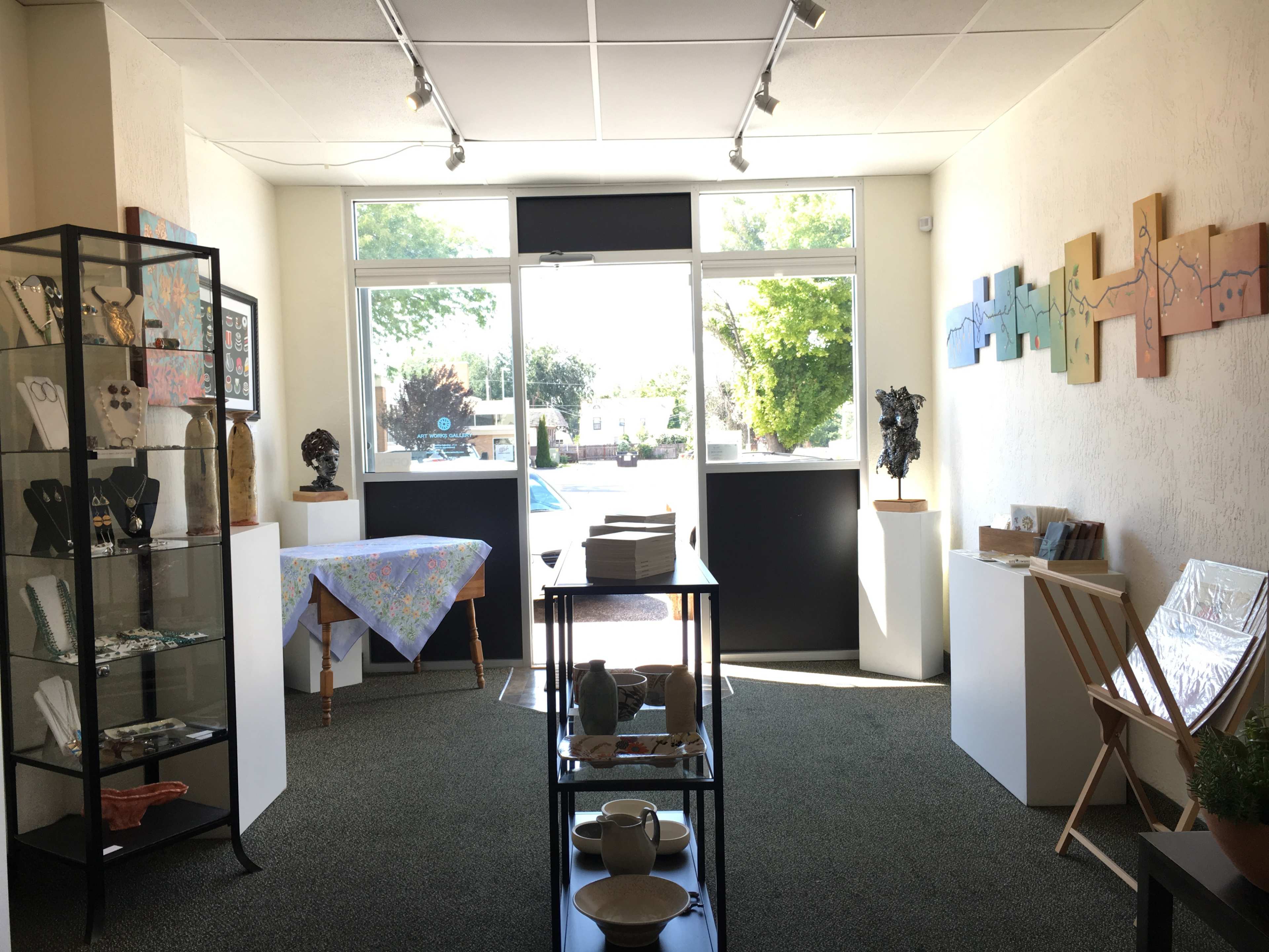 An art gallery interior with display cases and tables showcasing various artworks and crafts, illuminated by natural light from large windows.