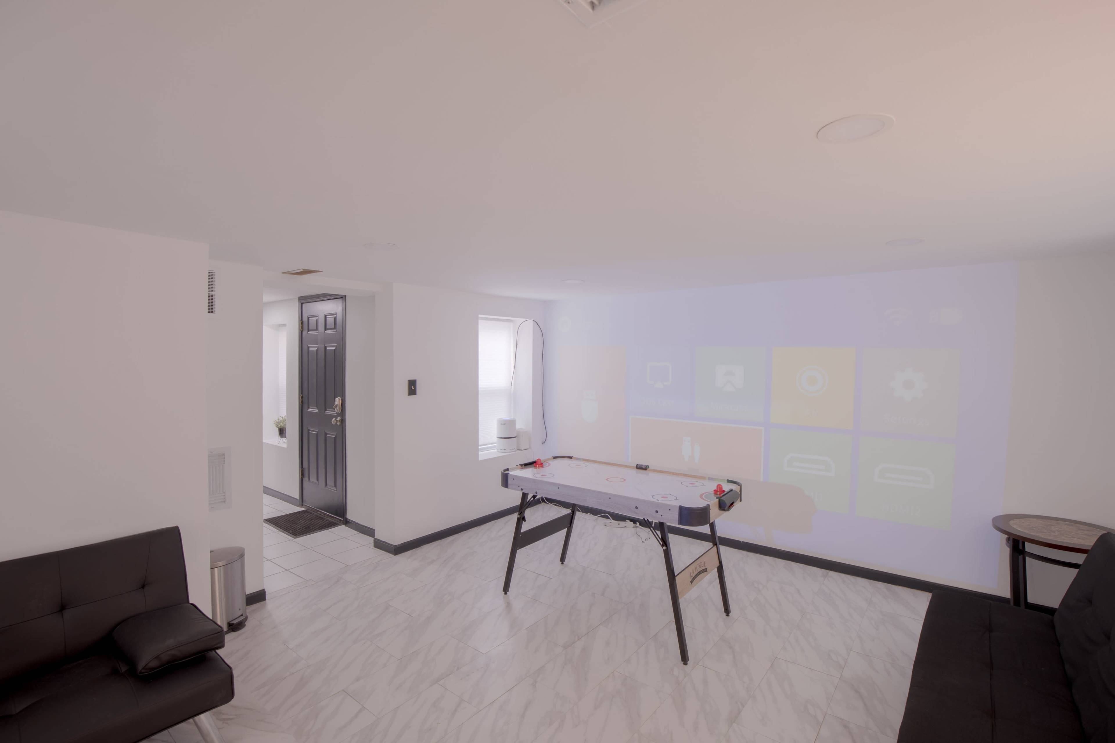A white room features a table soccer game in front of a wall displaying a projector screen with various interface icons.