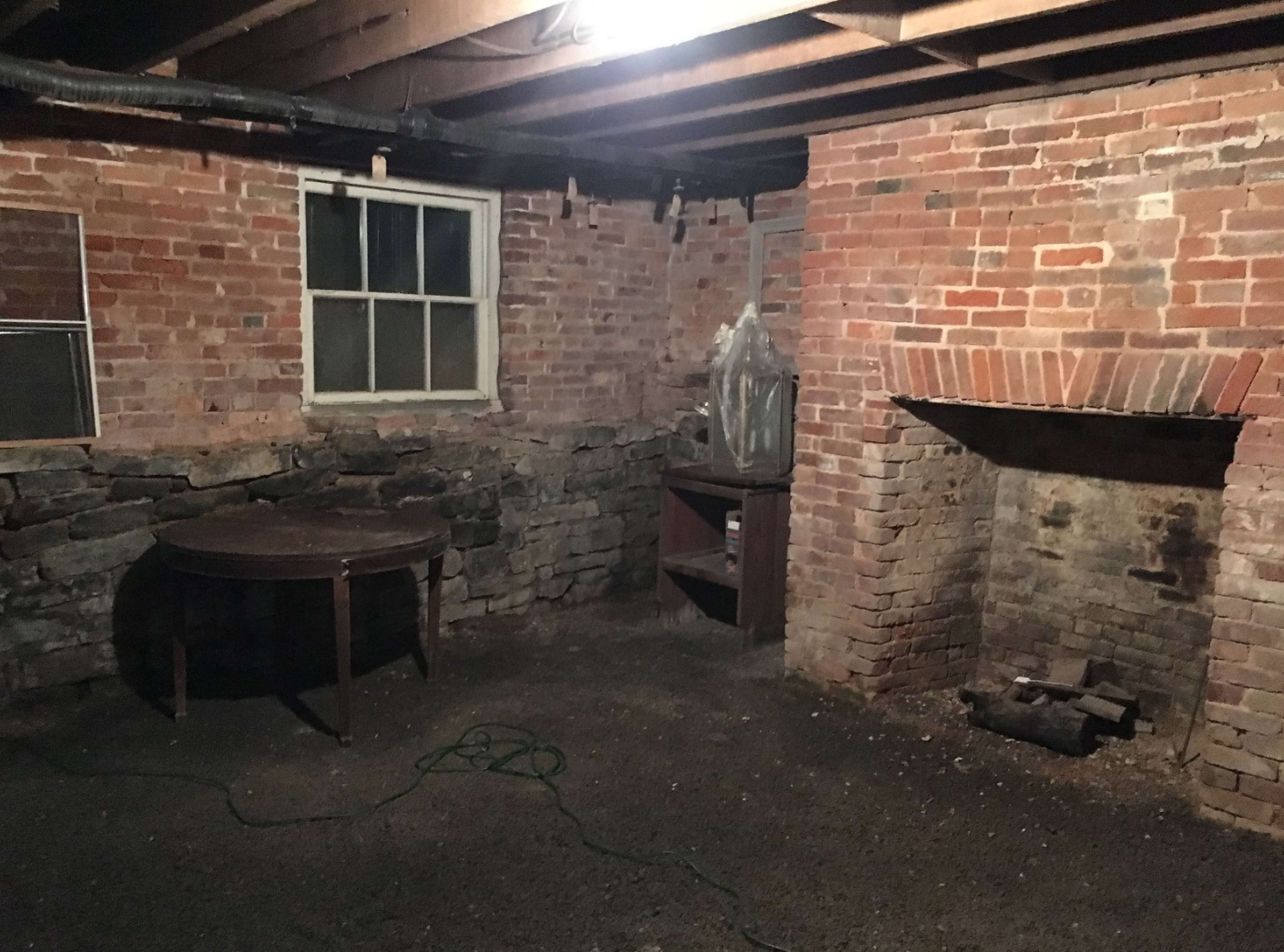 A dimly lit basement with exposed brick walls, a wooden table, a stone fireplace, and a window.