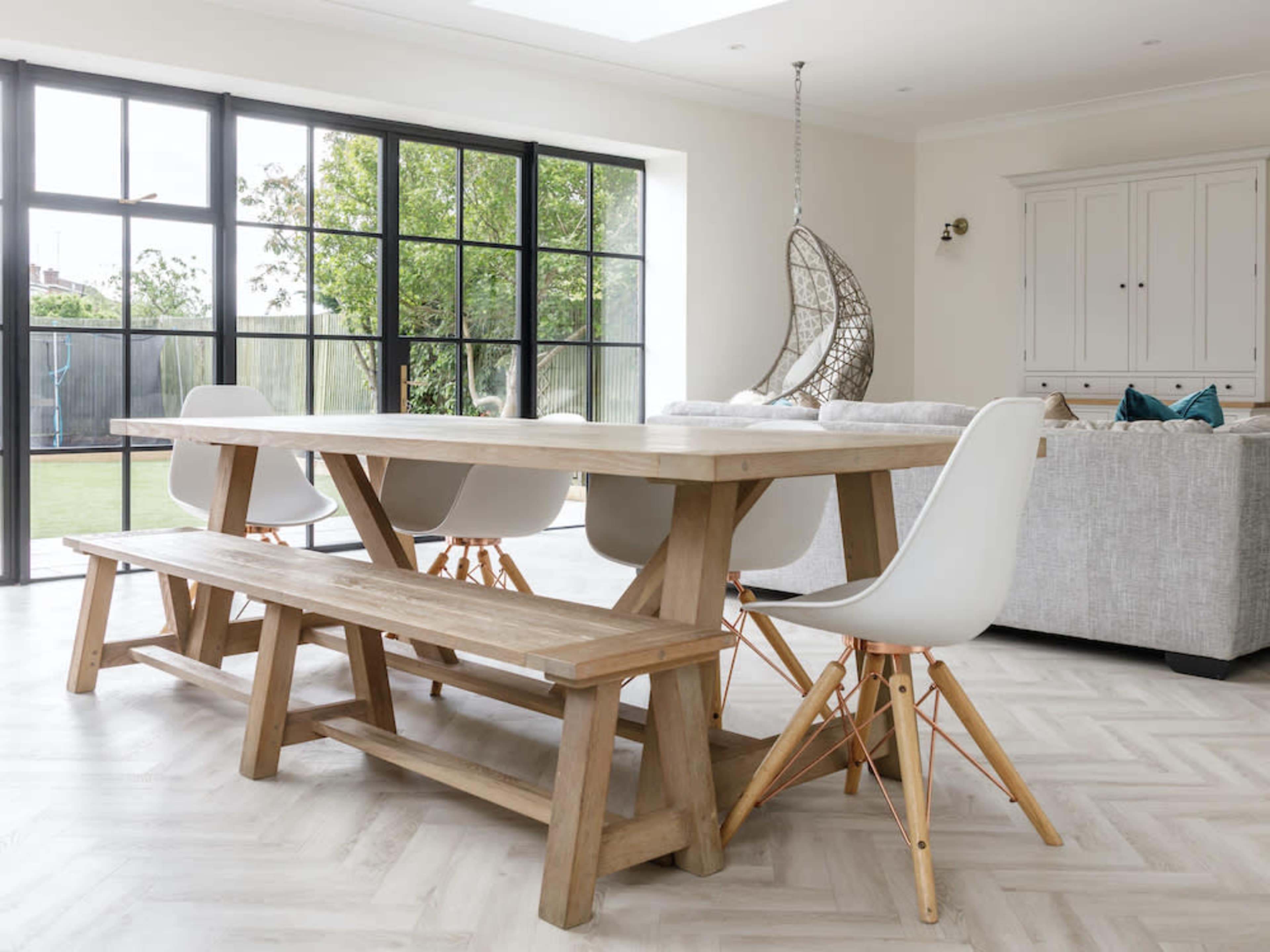 The image shows a modern dining area with a large wooden table, accompanied by white chairs, a bench, and a lounge area with a gray sofa and a hanging chair near large windows.