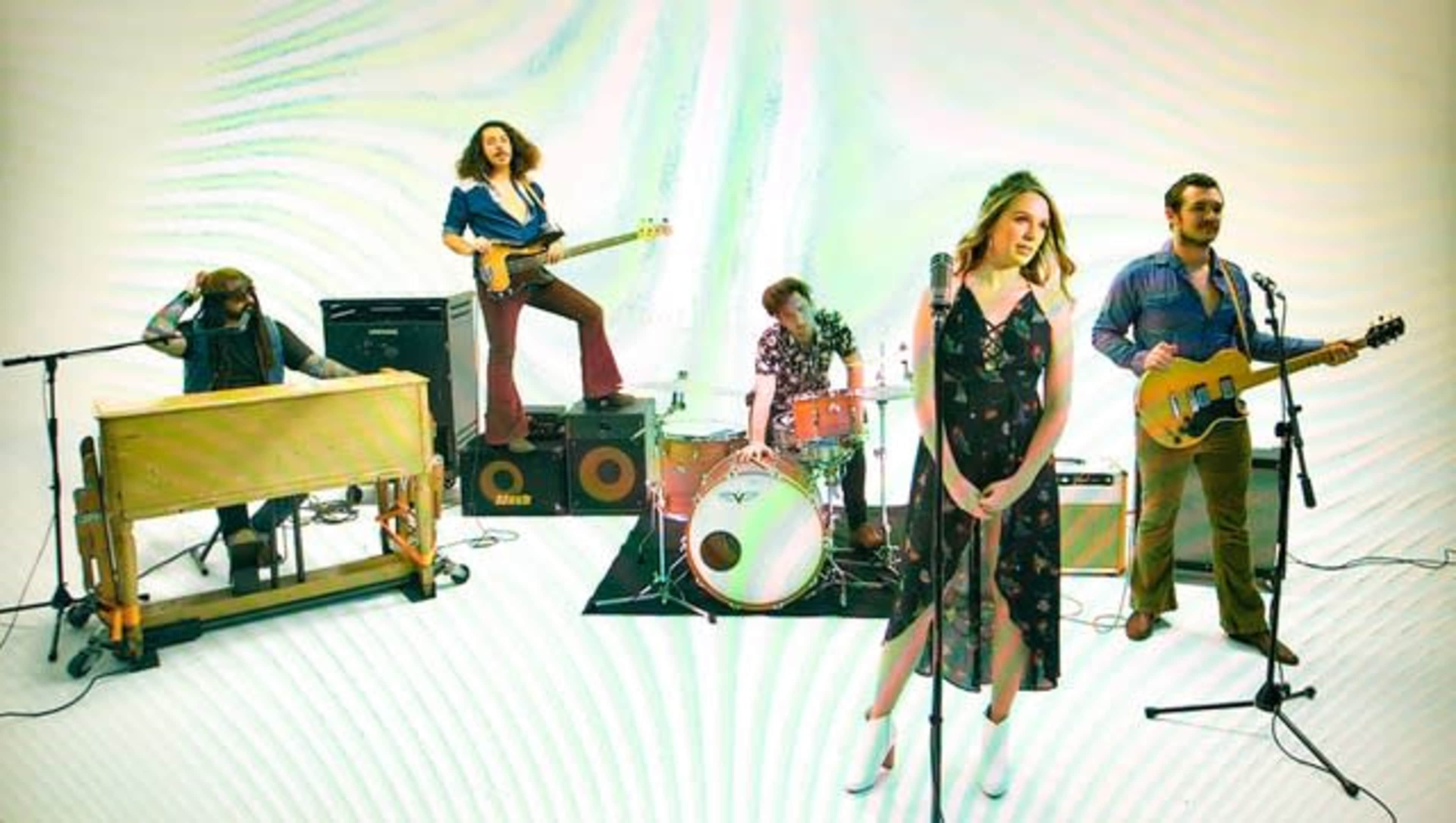 A band performs on a plain white backdrop, featuring a female vocalist, guitarists, a bassist, a drummer, and a keyboardist.