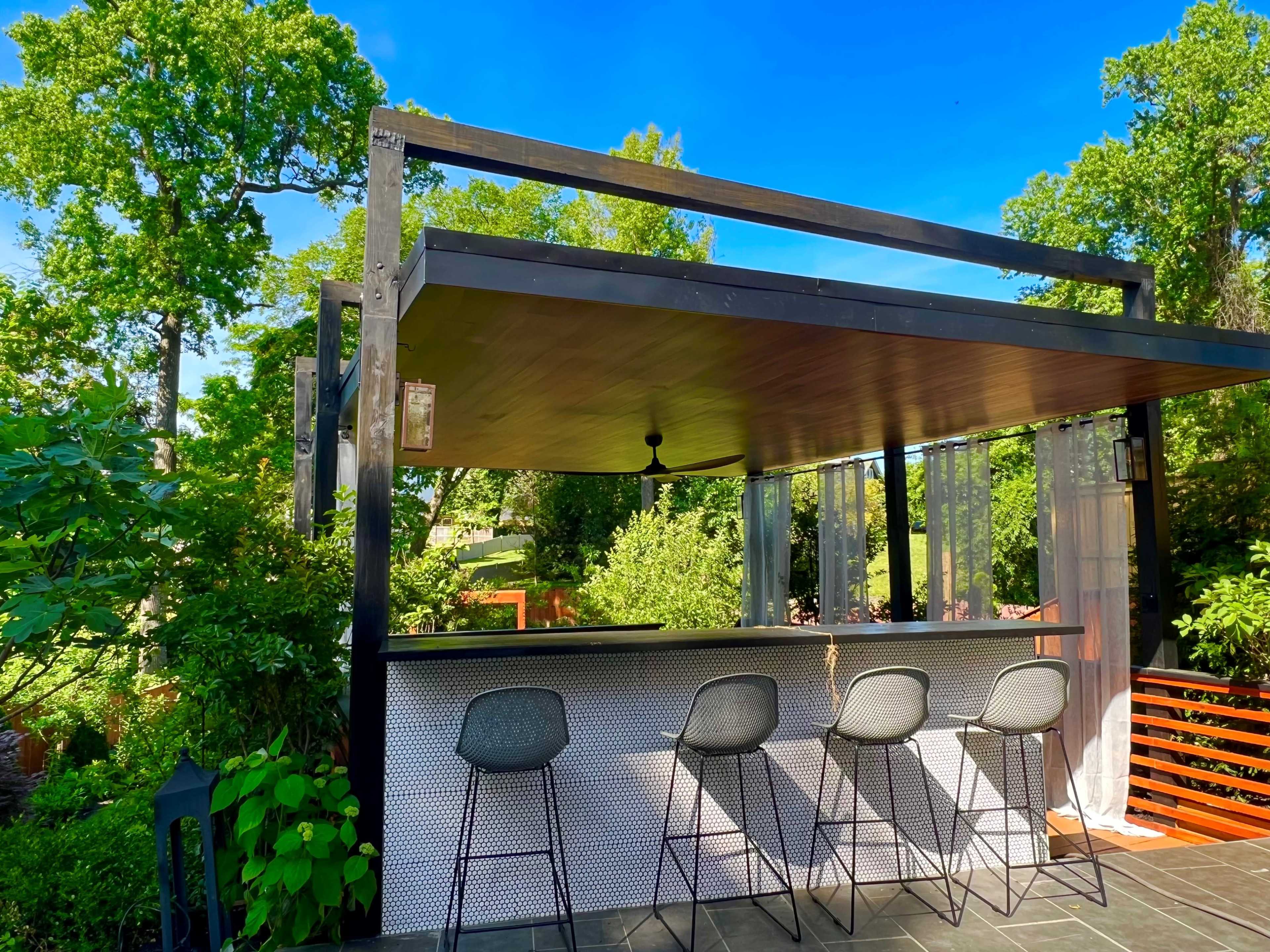 A modern outdoor bar area with four high stools, set under a roof supported by a sleek black frame, surrounded by greenery.
