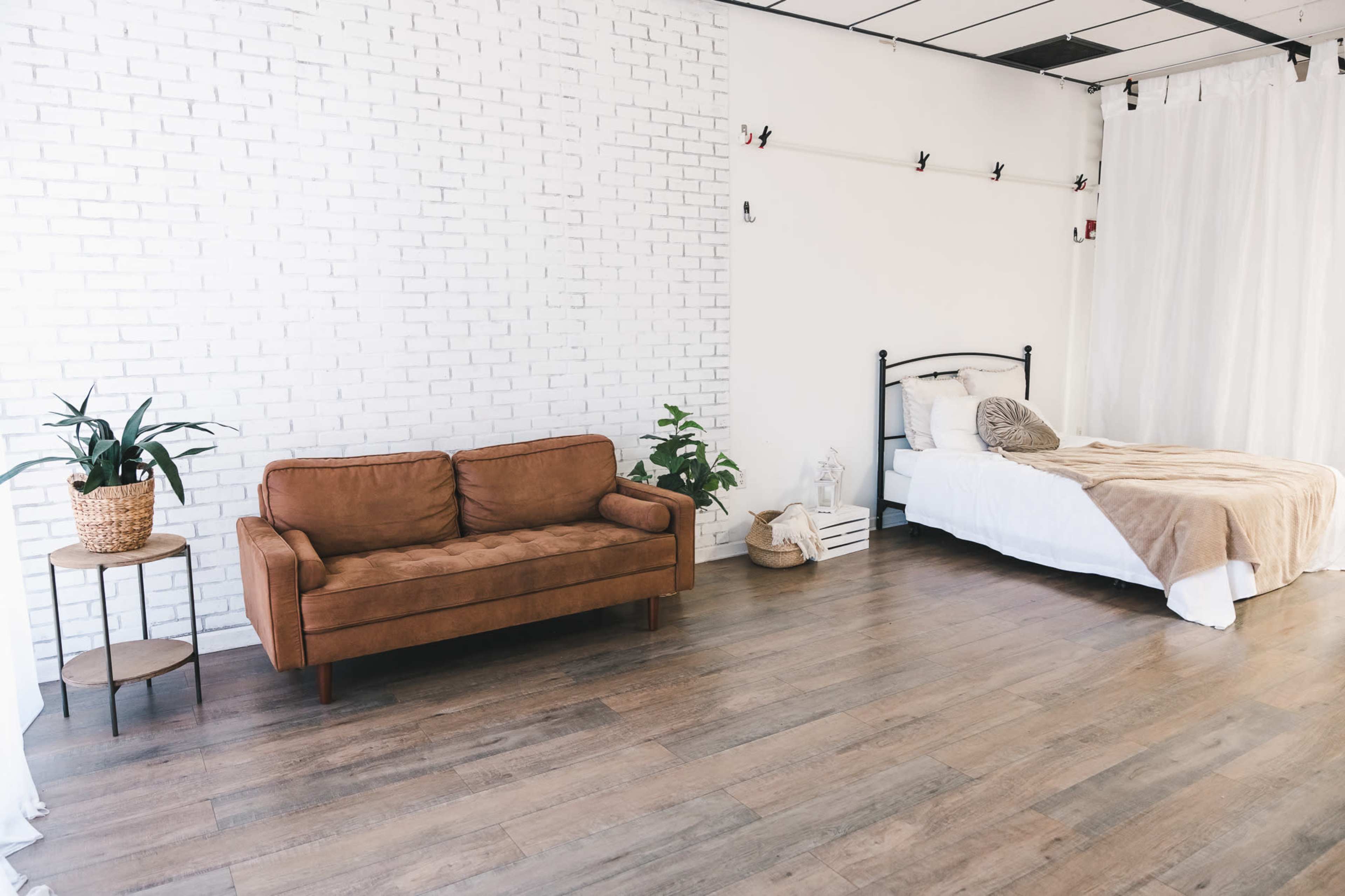A minimalist room features a brown couch, a bed with white bedding, and light-colored walls, with plants and a side table included in the decor.