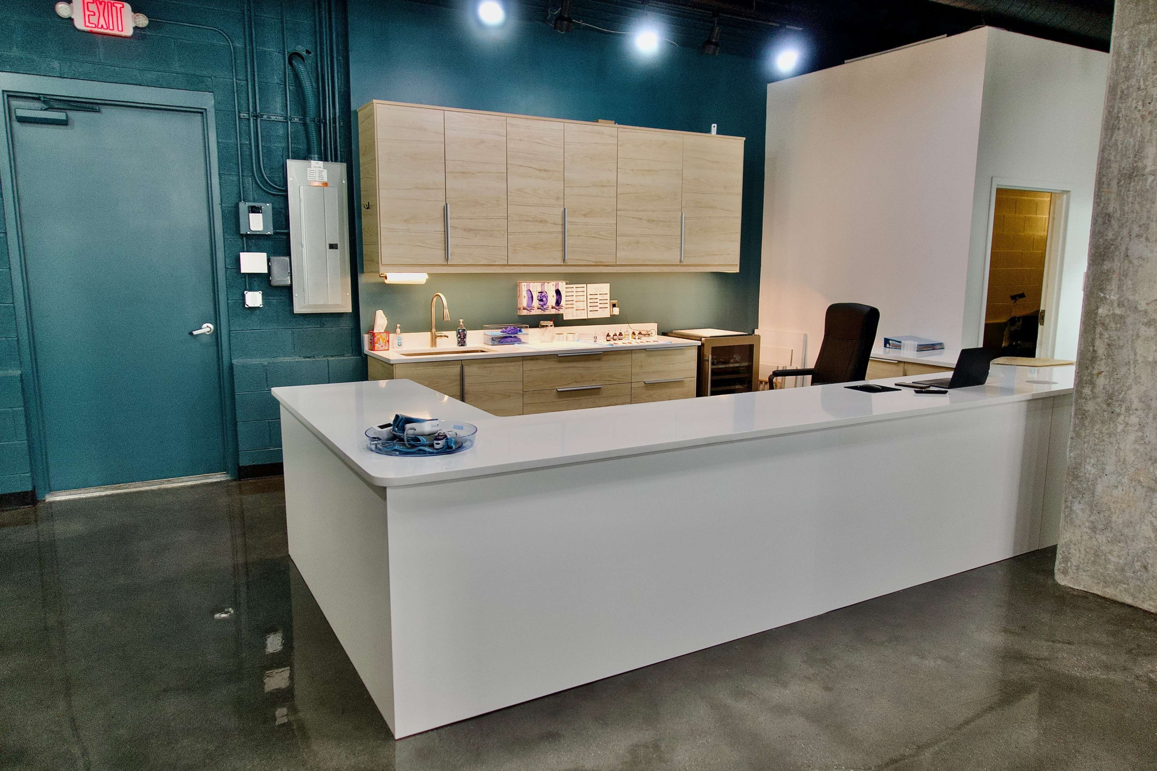 The image shows a modern reception area with a white front desk, wooden cabinetry, and a dark teal wall.