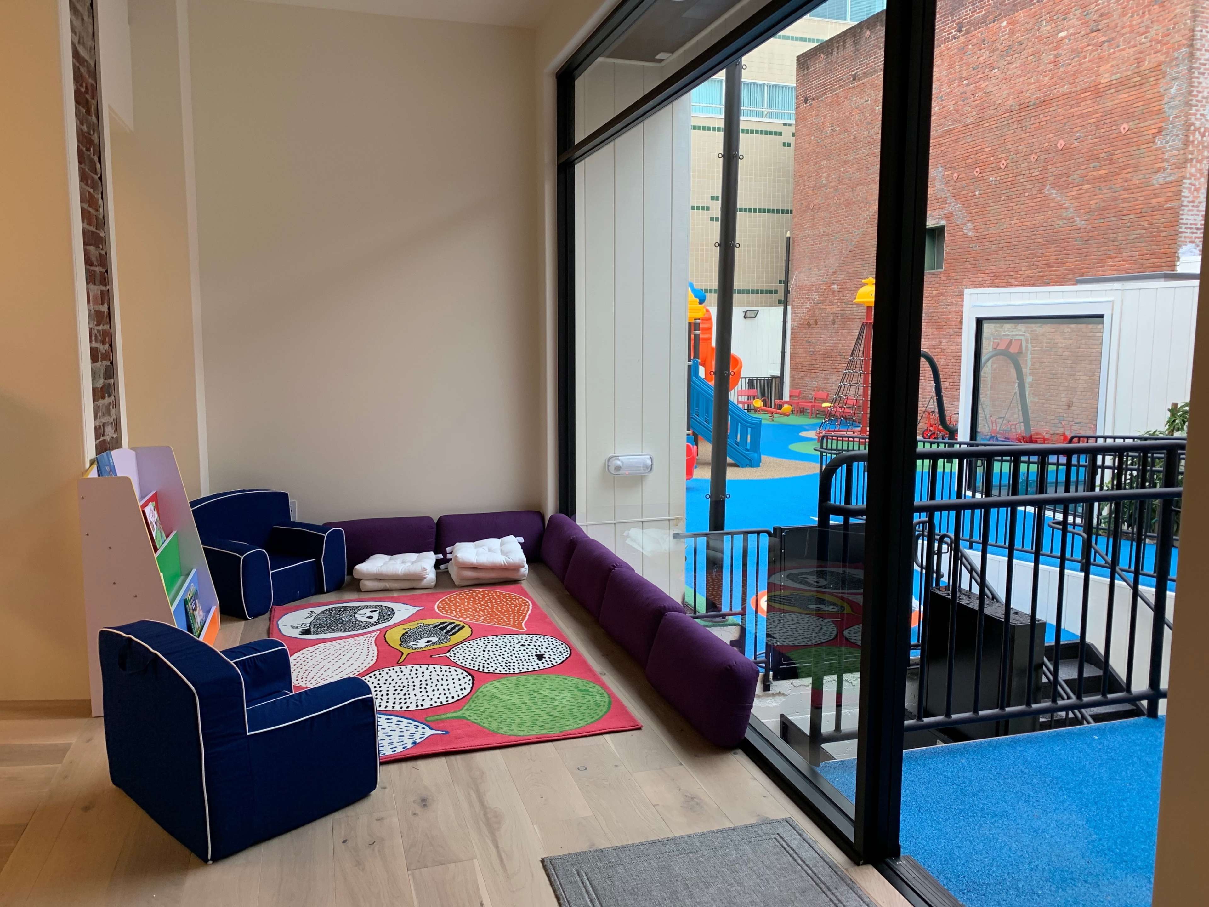 A small indoor play area features a colorful rug, low seating, and a large window overlooking an outdoor playground with toys and play structures.