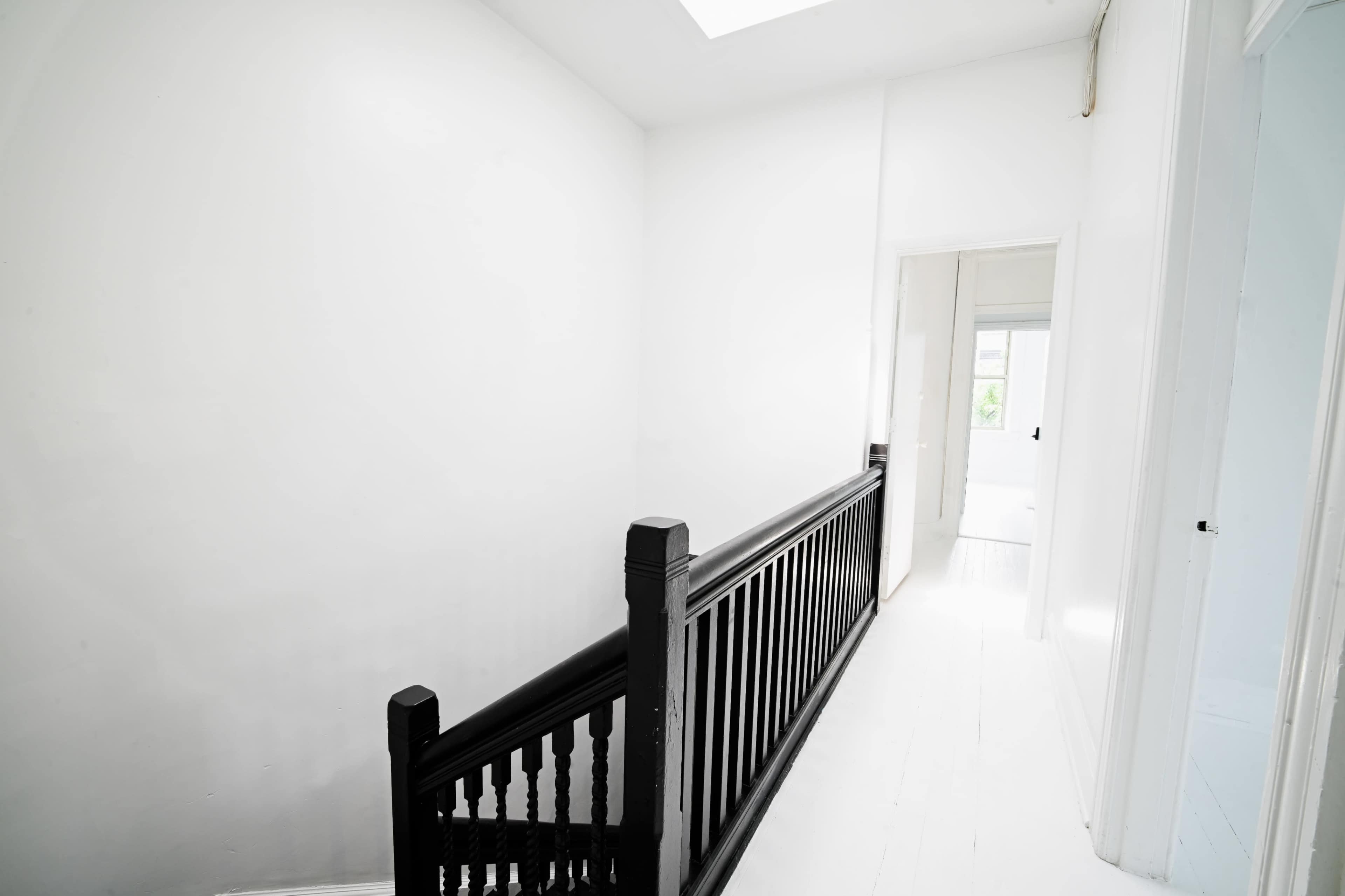 The image shows a narrow hallway with white walls, a black wooden railing, and a doorway leading to a light-filled room at the end.