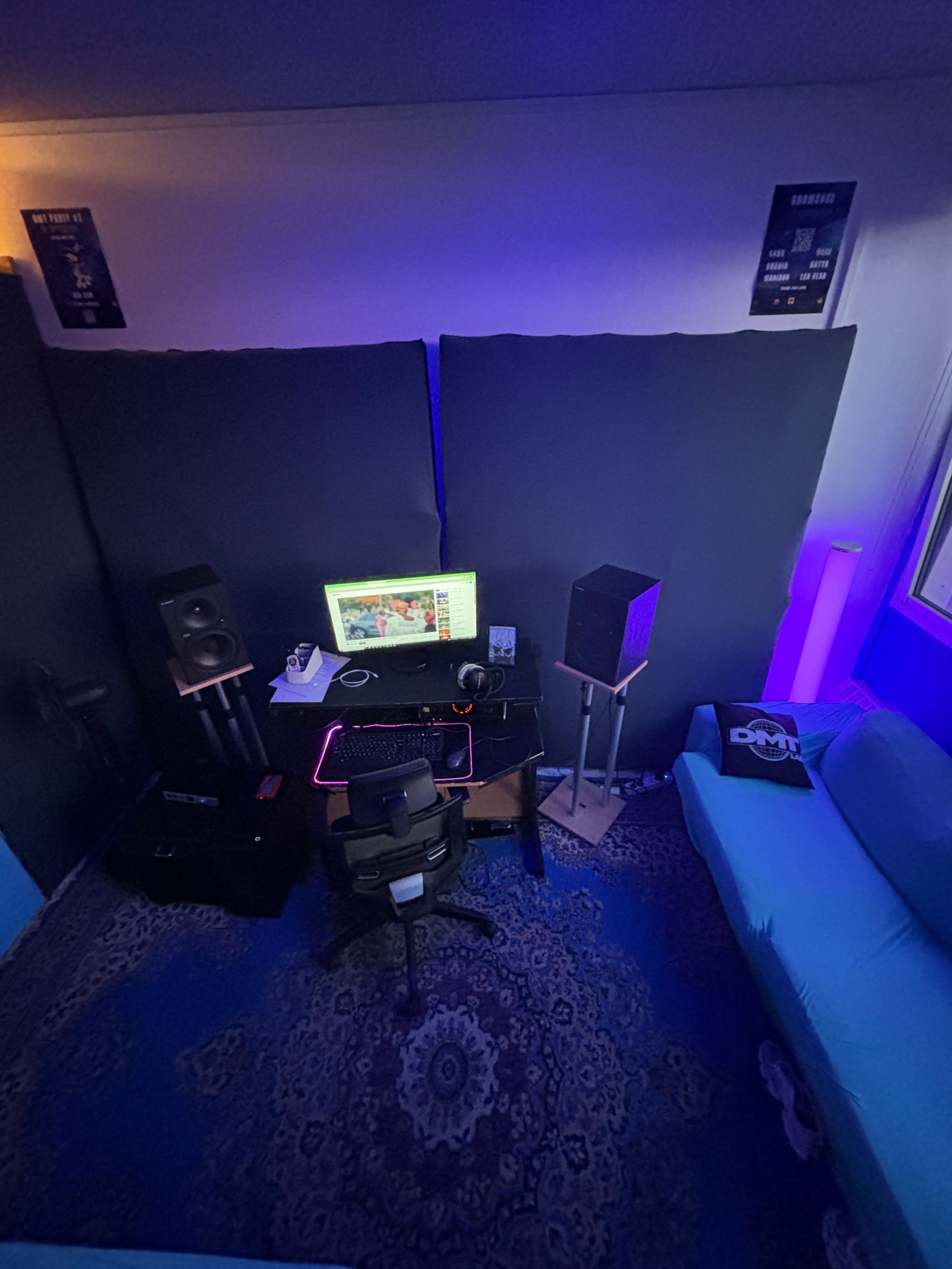 A small room features a desk with a computer, speakers, and a chair, surrounded by soundproofing panels and illuminated by soft purple lighting.