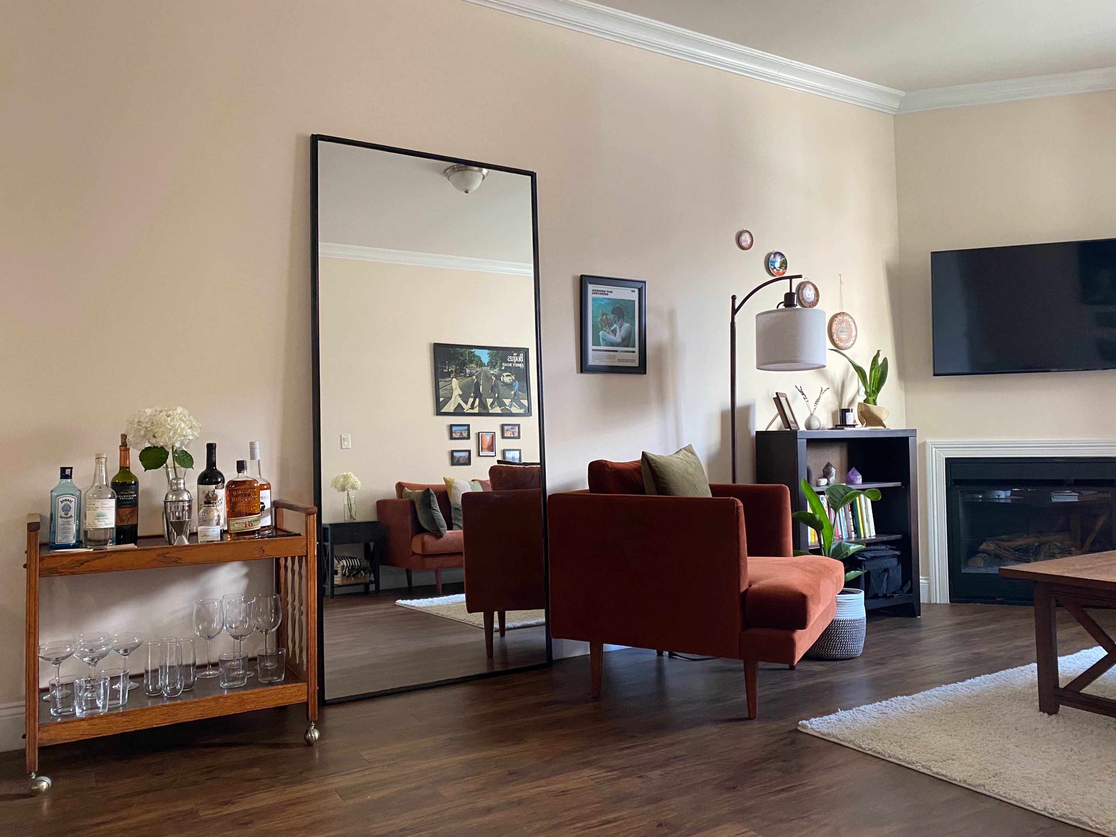 A living room features a large mirror, a bar cart with glassware and bottles, a red couch, a modern lamp, and a wall-mounted TV.