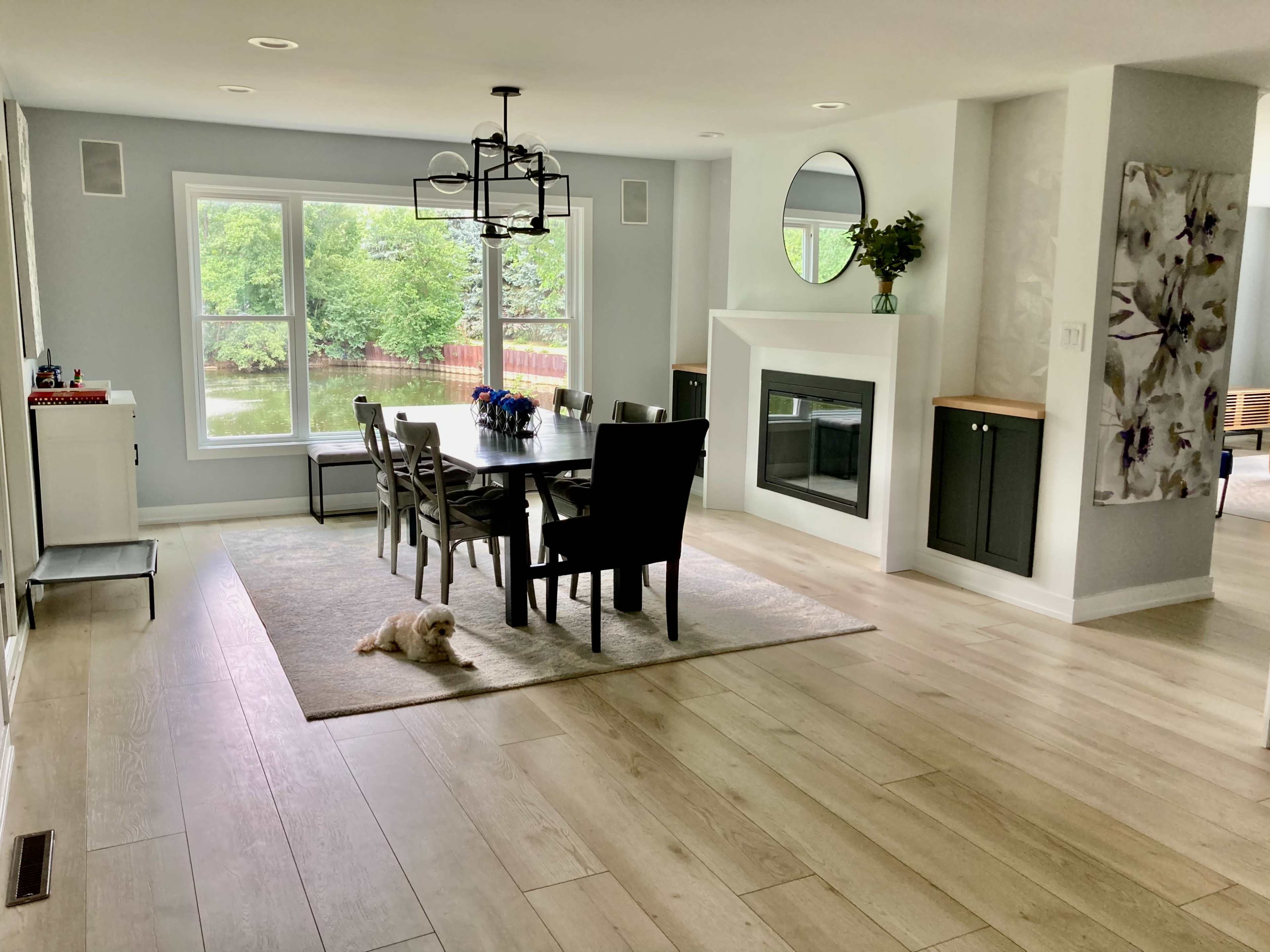 The interior of a modern dining area features a large table surrounded by chairs, a fireplace, and a window overlooking a wooded area.