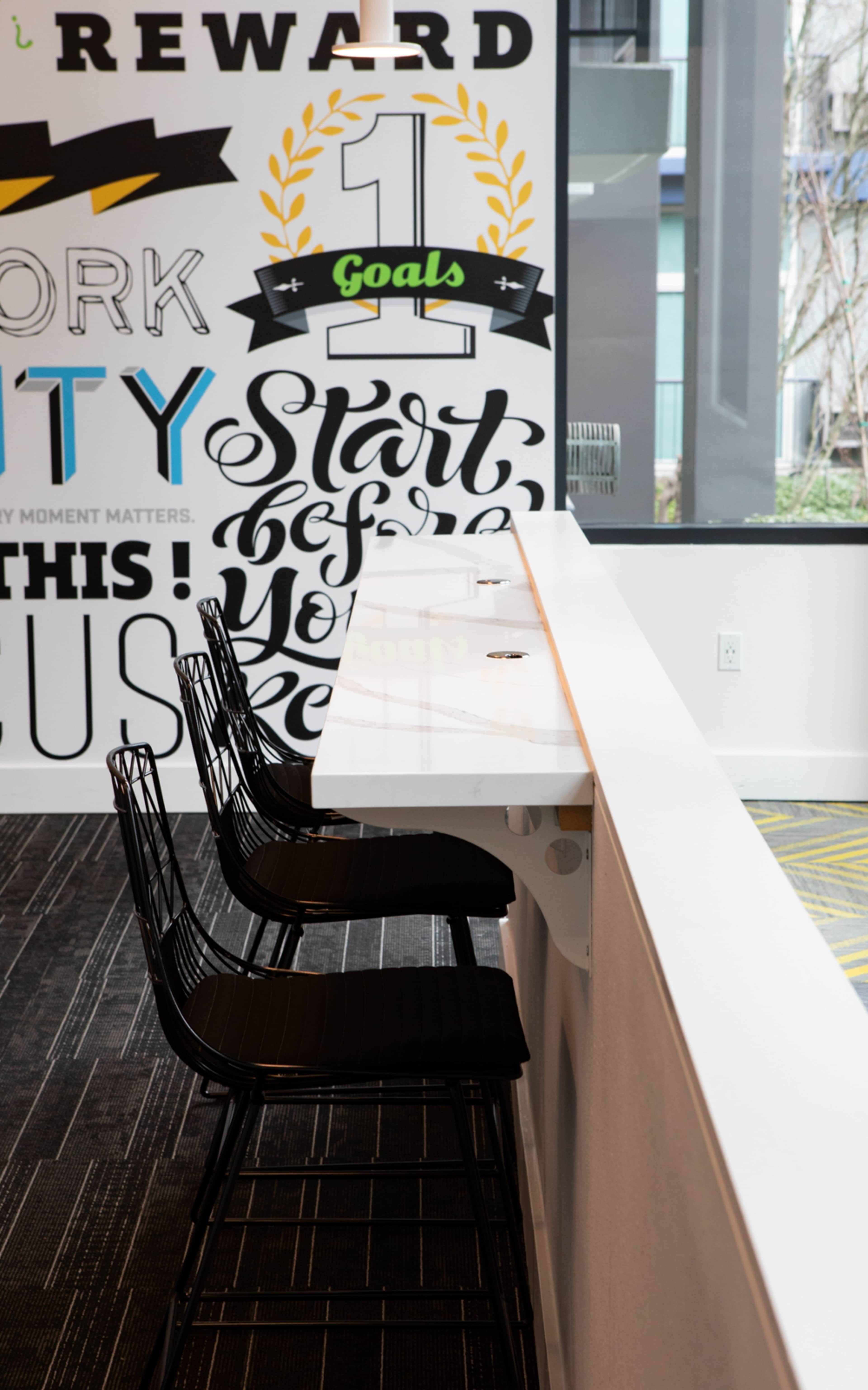 A row of black wire chairs is positioned at a long white marble countertop against a wall featuring motivational text and graphics.