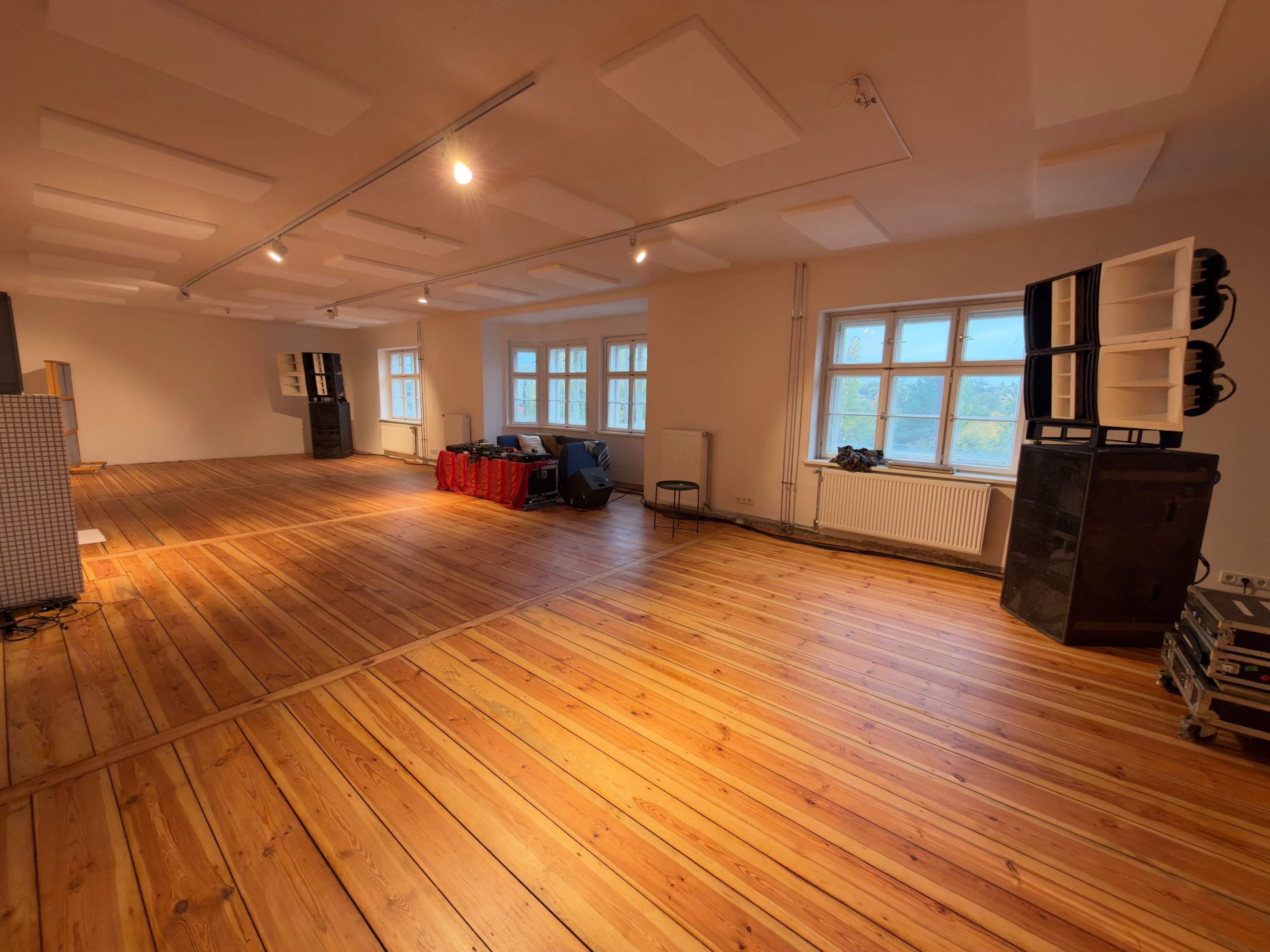 The image shows a spacious, empty room with wooden flooring, large windows, and stacked equipment against the walls.