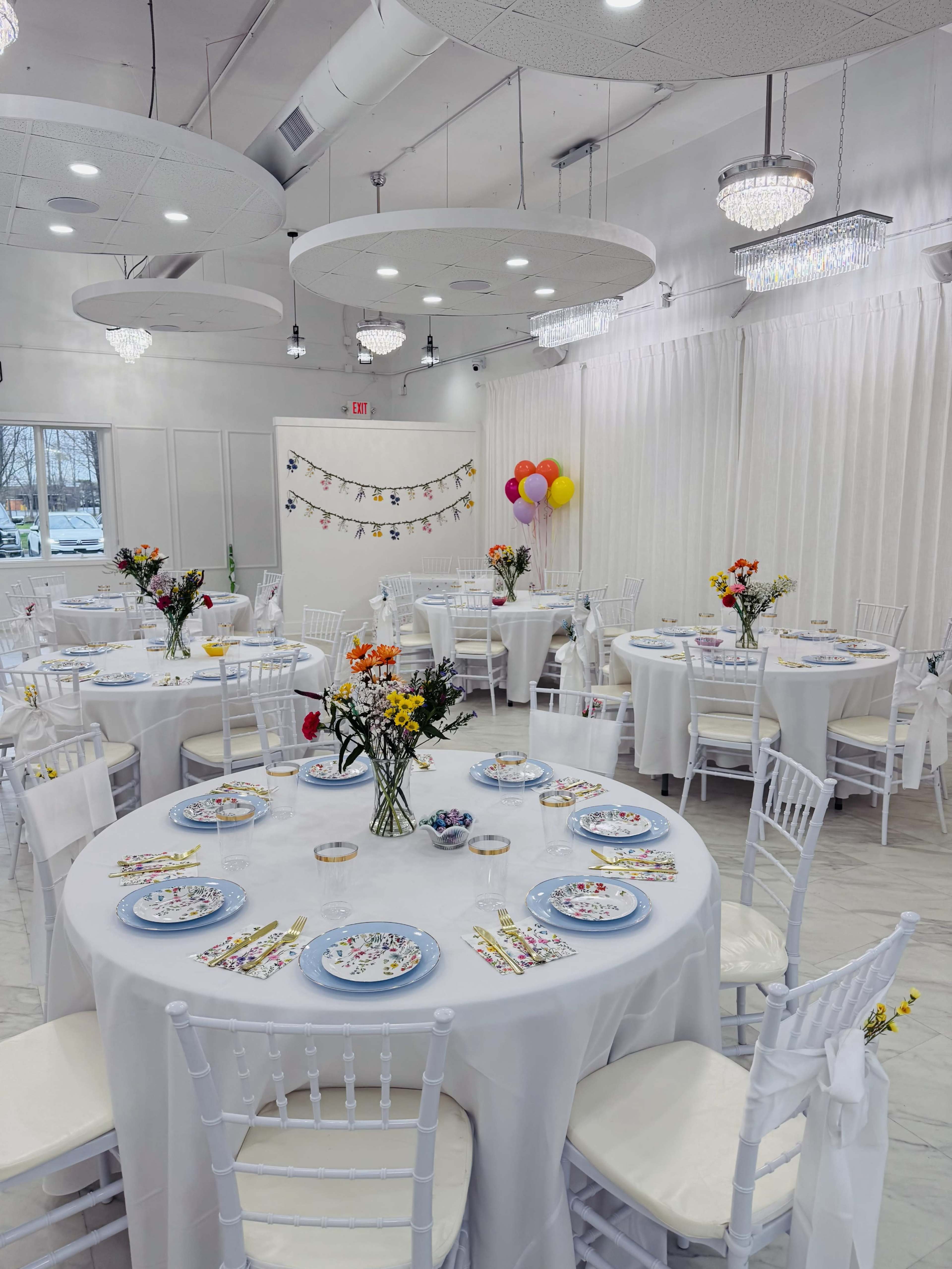 The image shows a bright banquet hall set with circular tables adorned with white tablecloths, plates, and floral centerpieces, along with decorations on the walls.