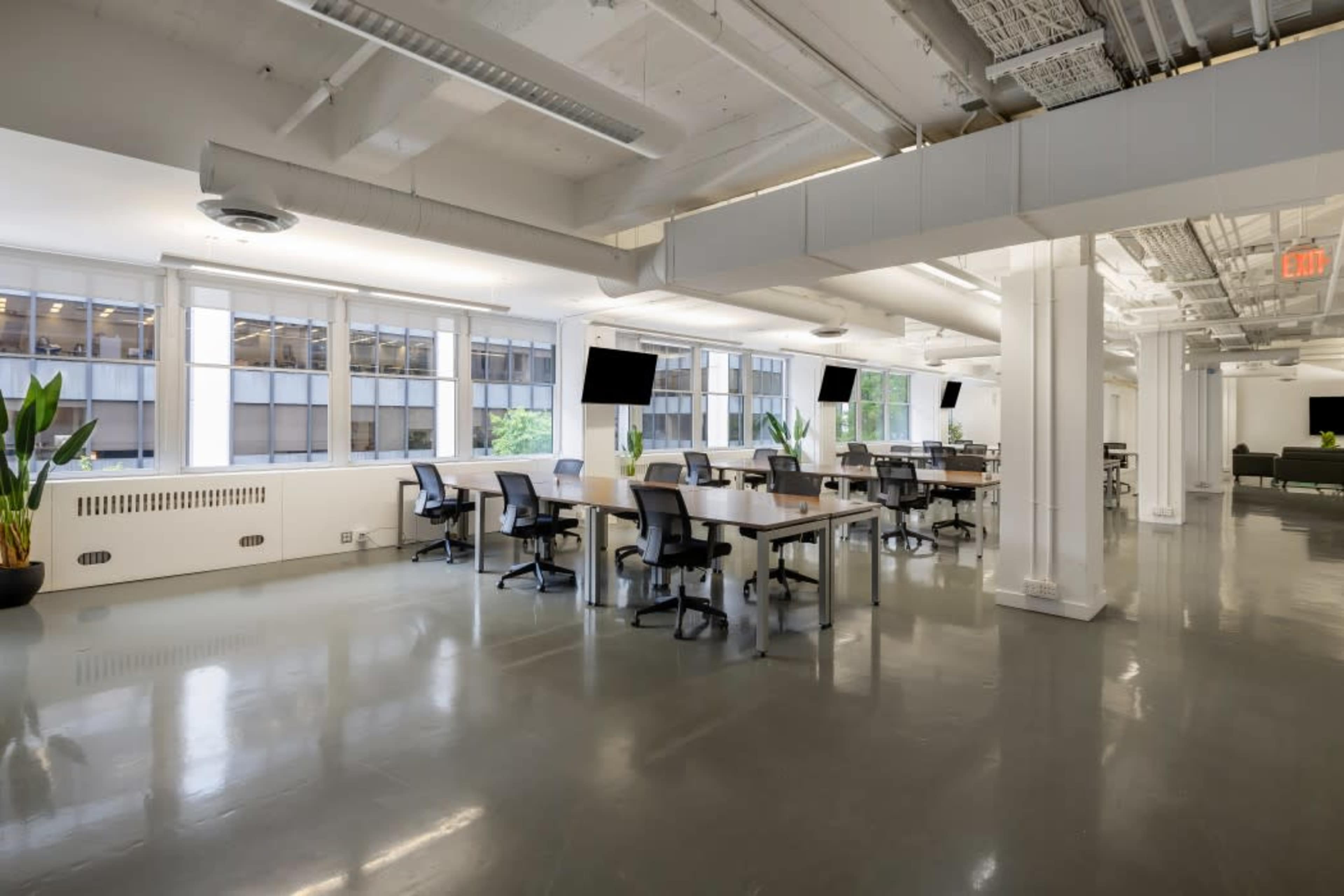 A spacious, modern office interior featuring multiple desks with chairs, large windows, and plant decor.