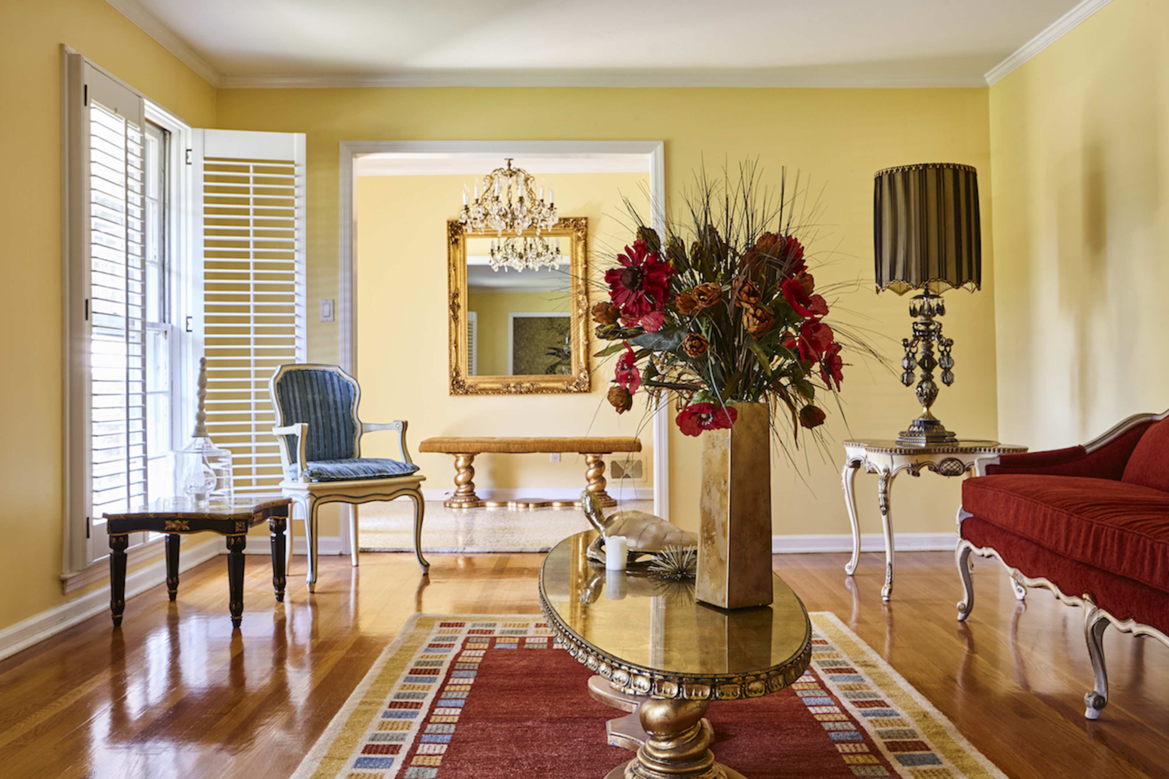 A brightly colored living room features a red sofa, a wooden coffee table, a decorative lamp, and a vase of flowers on a patterned rug.