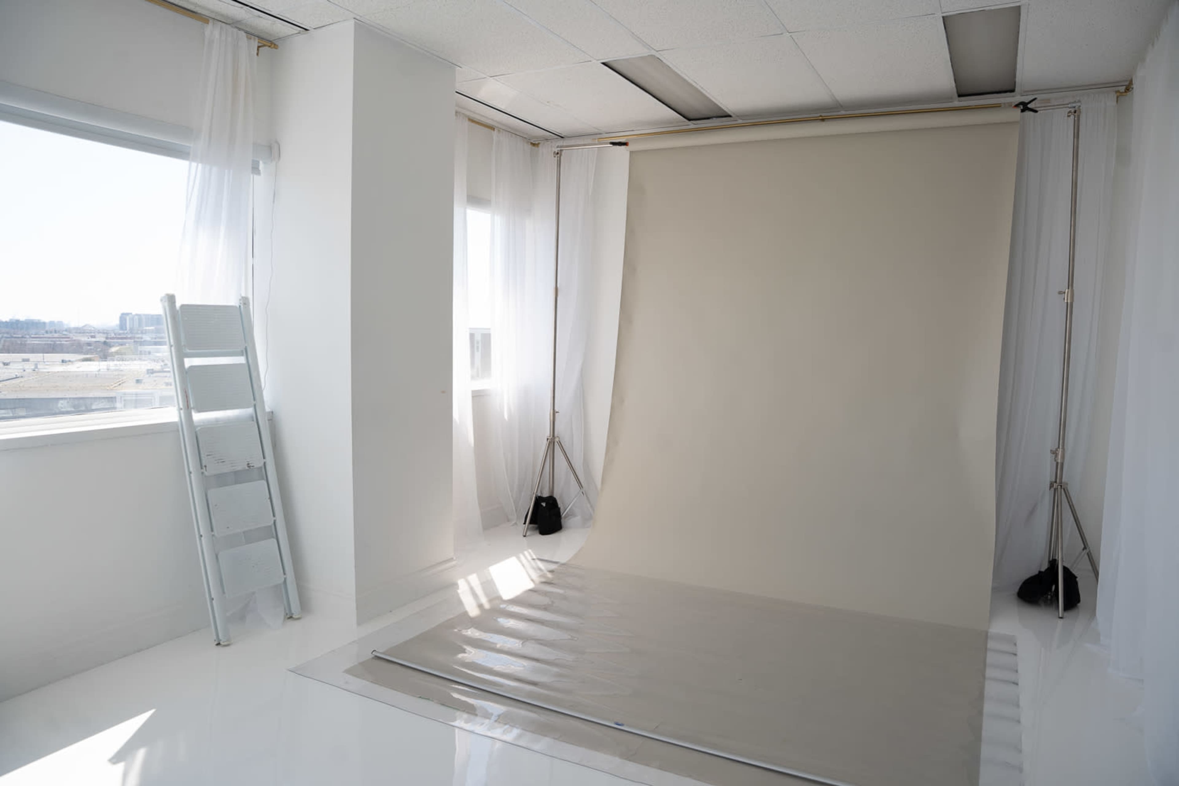 The image shows a photography studio with a large blank backdrop, a shiny floor, and a ladder leaning against the wall near a window with sheer curtains.