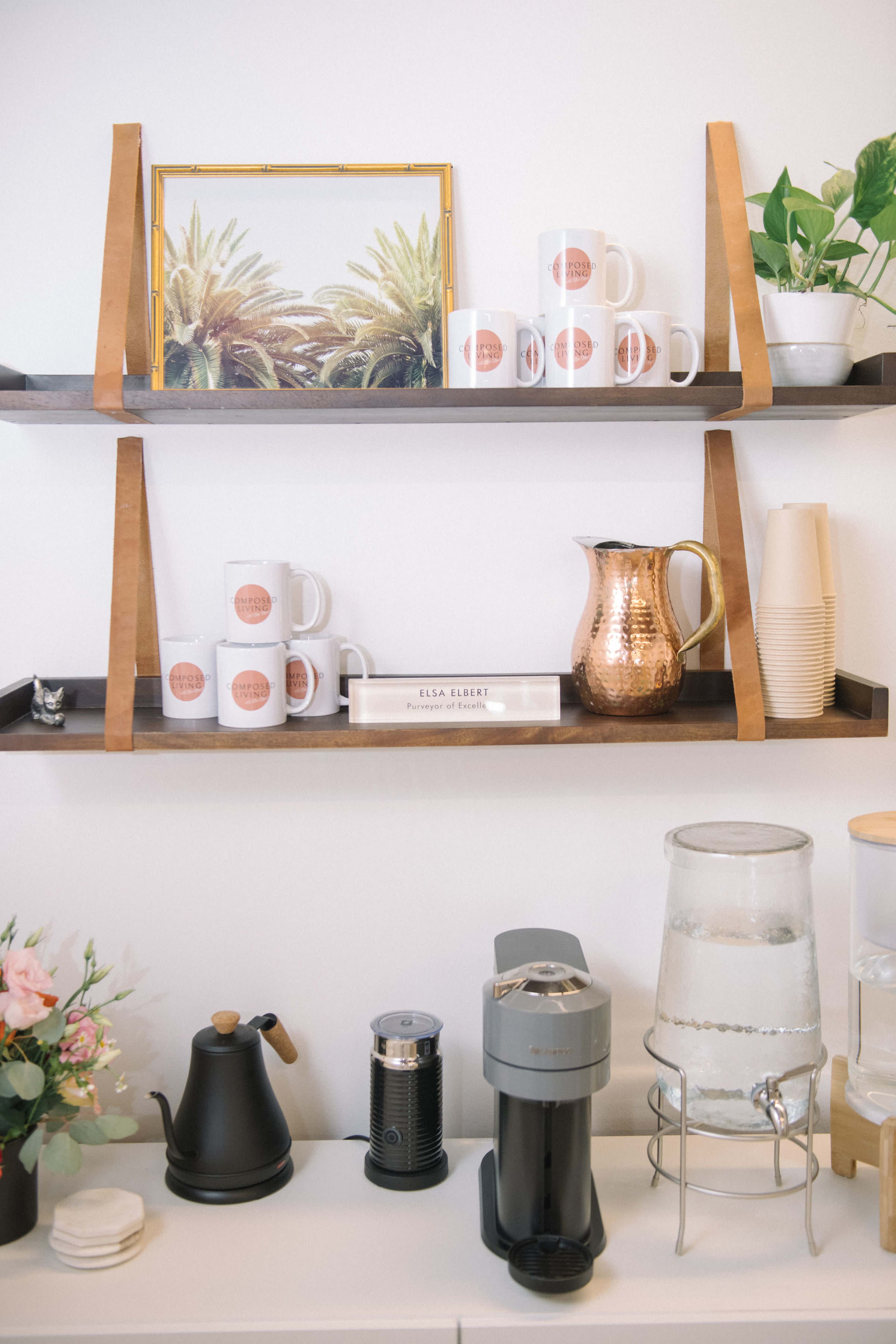 A wooden shelf displaying mugs, a decorative frame, a copper pitcher, and a coffee maker, with a water dispenser and a kettle on a counter below.
