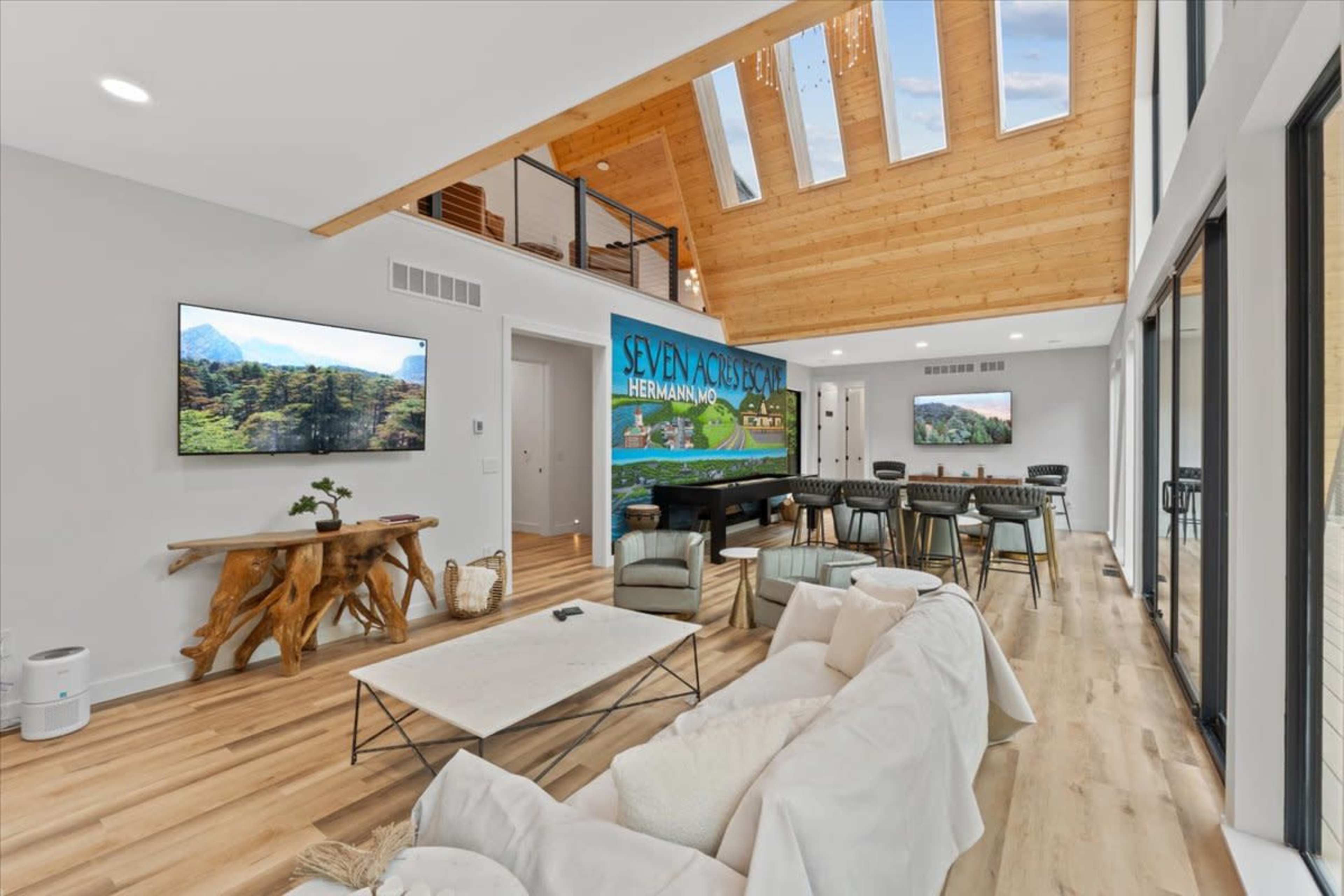 The image shows a modern living space with a vaulted wooden ceiling, large windows, and an open layout featuring a couch, dining area, and two wall-mounted televisions.