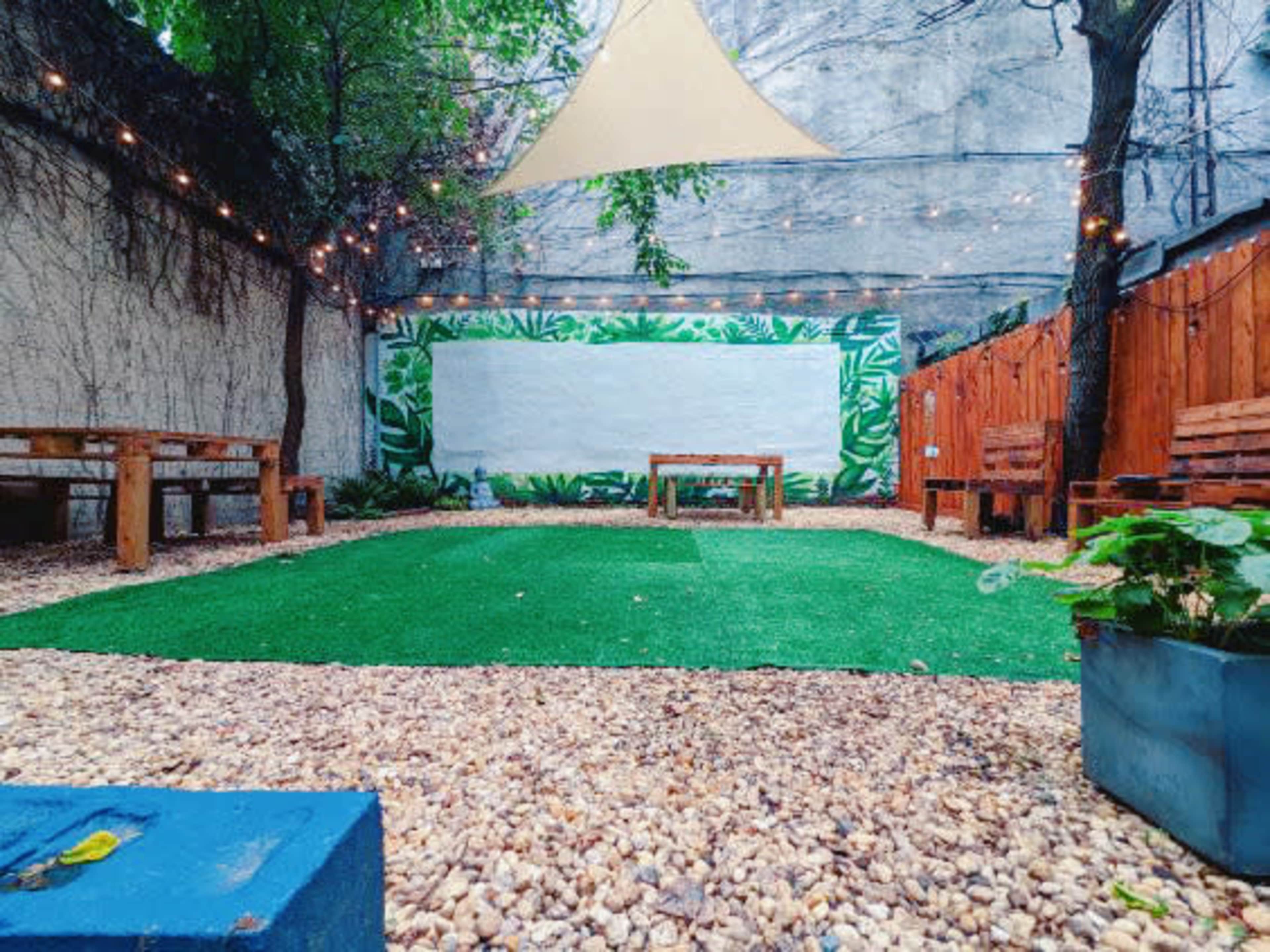 The image shows a garden space featuring wooden seating, a central table, decorative plants, and a grassy area surrounded by a rocky landscape and a mural.