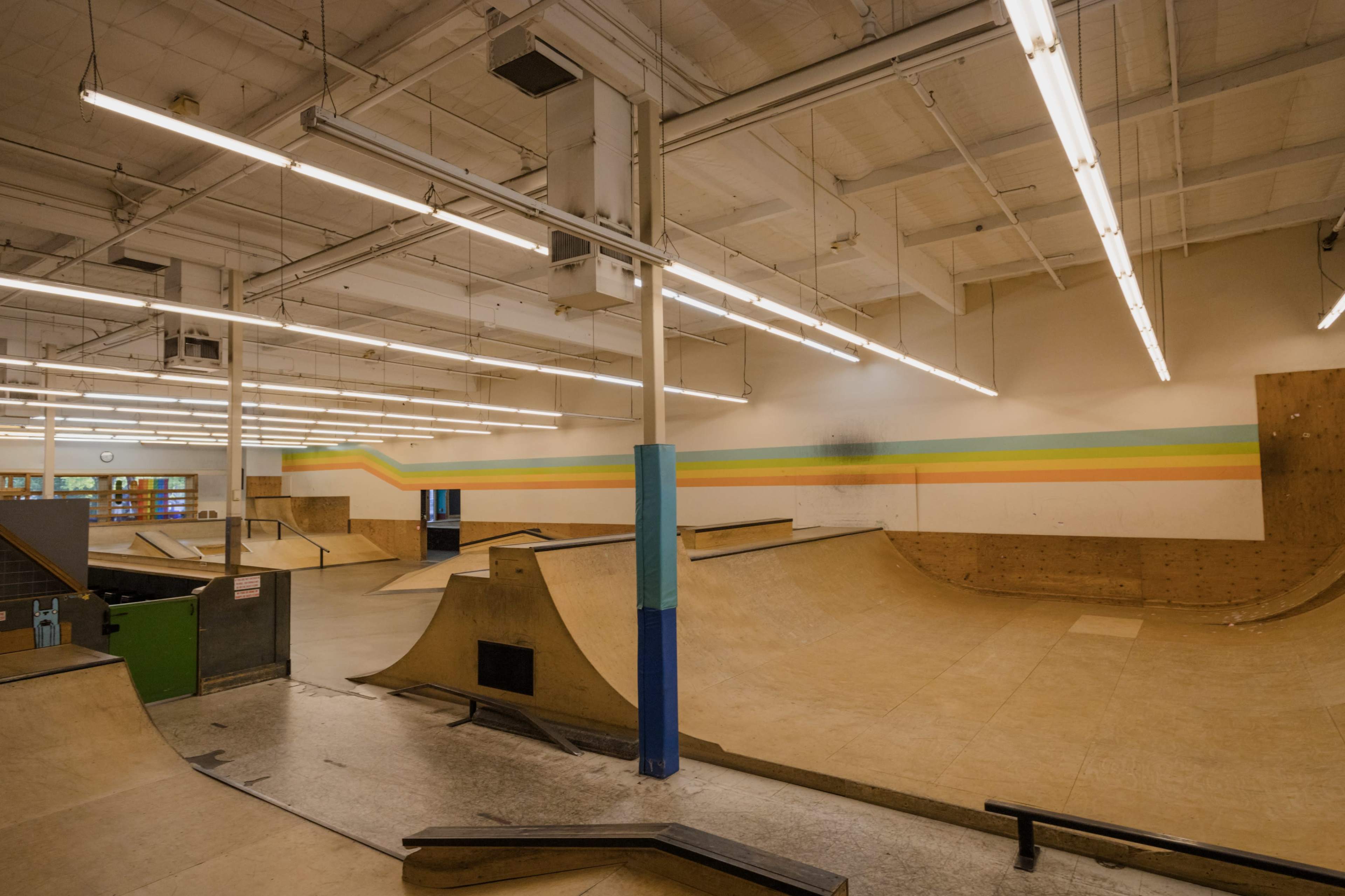 The image shows an indoor skate park with wooden ramps and a smooth concrete floor beneath bright overhead lighting.
