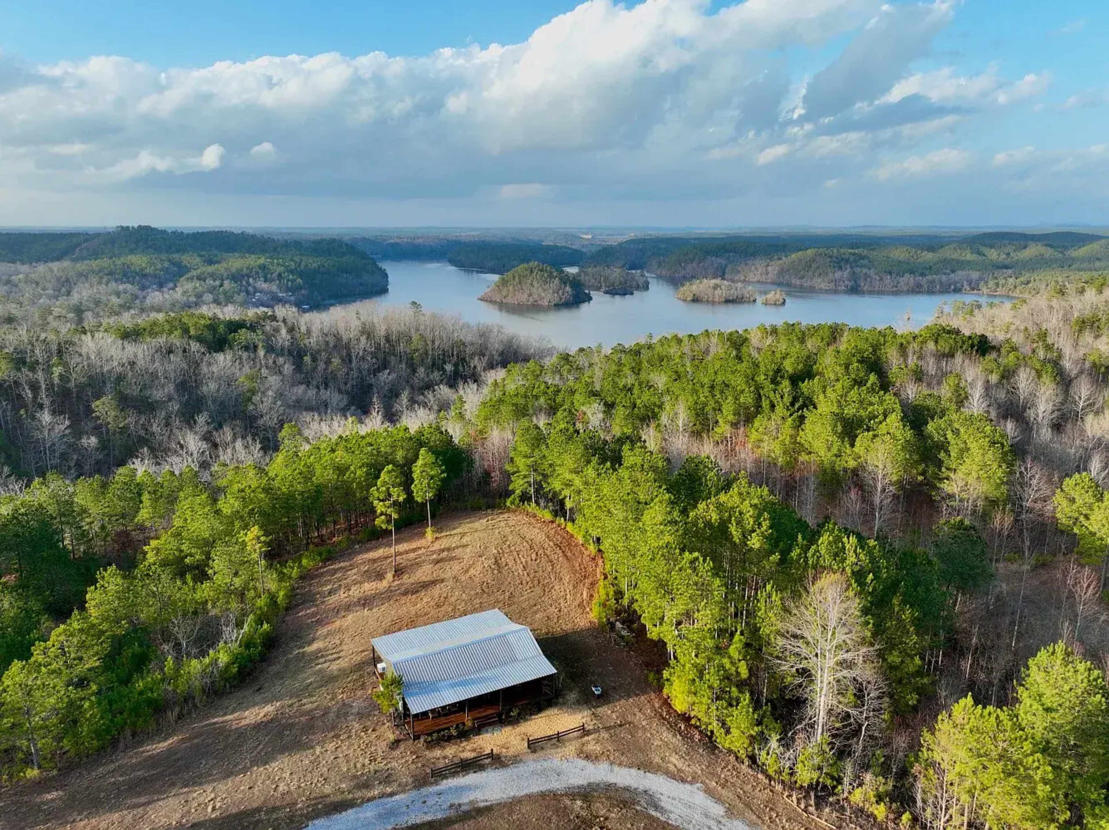 A wooden cabin with a metal roof is situated near a lake, surrounded by patches of forest and hilly terrain.
