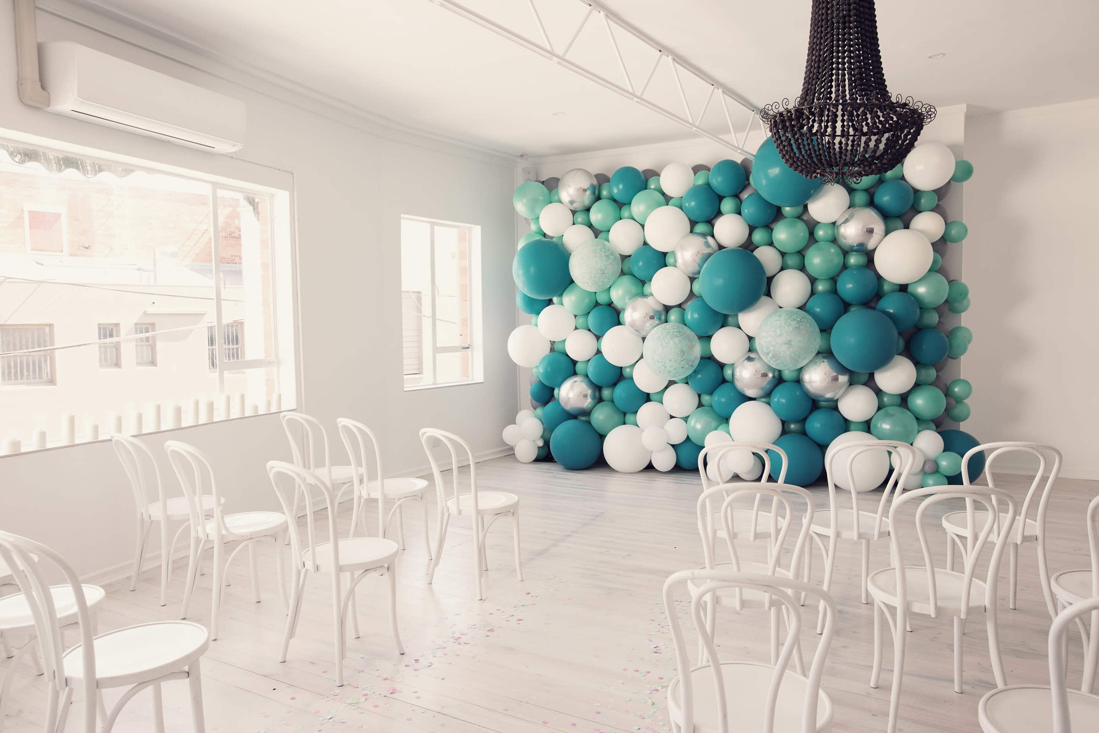 A room with a decorative wall of balloons in shades of teal and white, accompanied by several white chairs facing the wall.