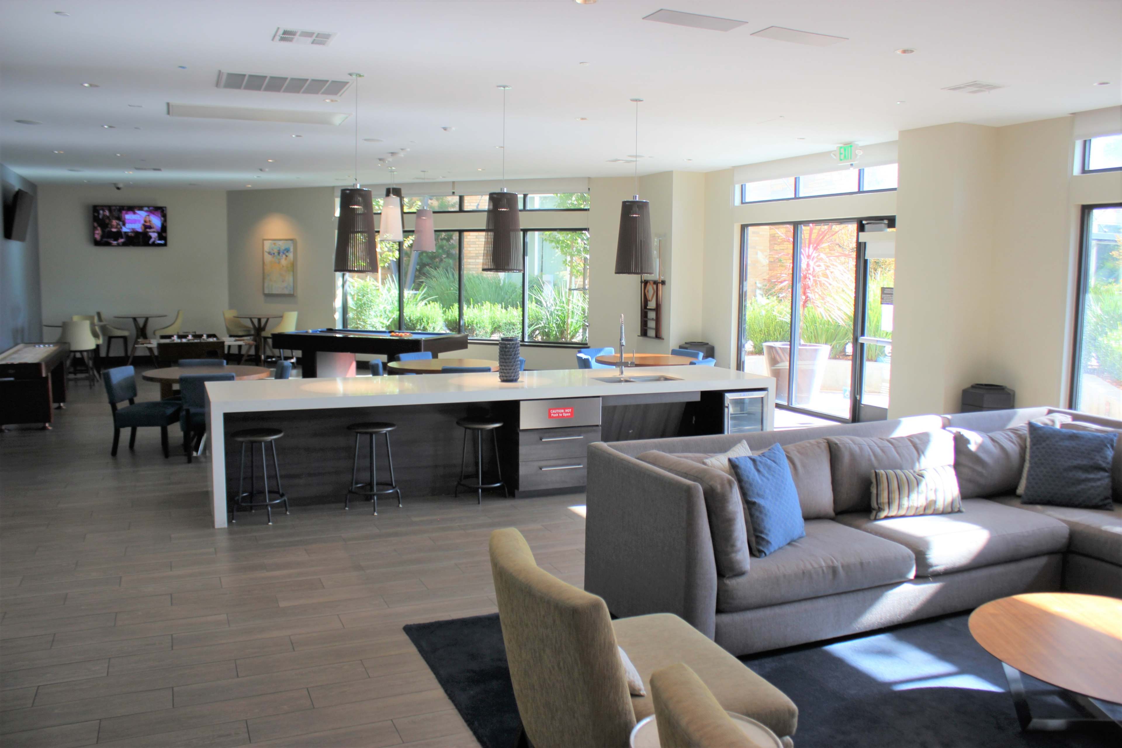 A modern lounge area featuring a kitchen bar, seating with gray sofas, and large windows illuminating the space.