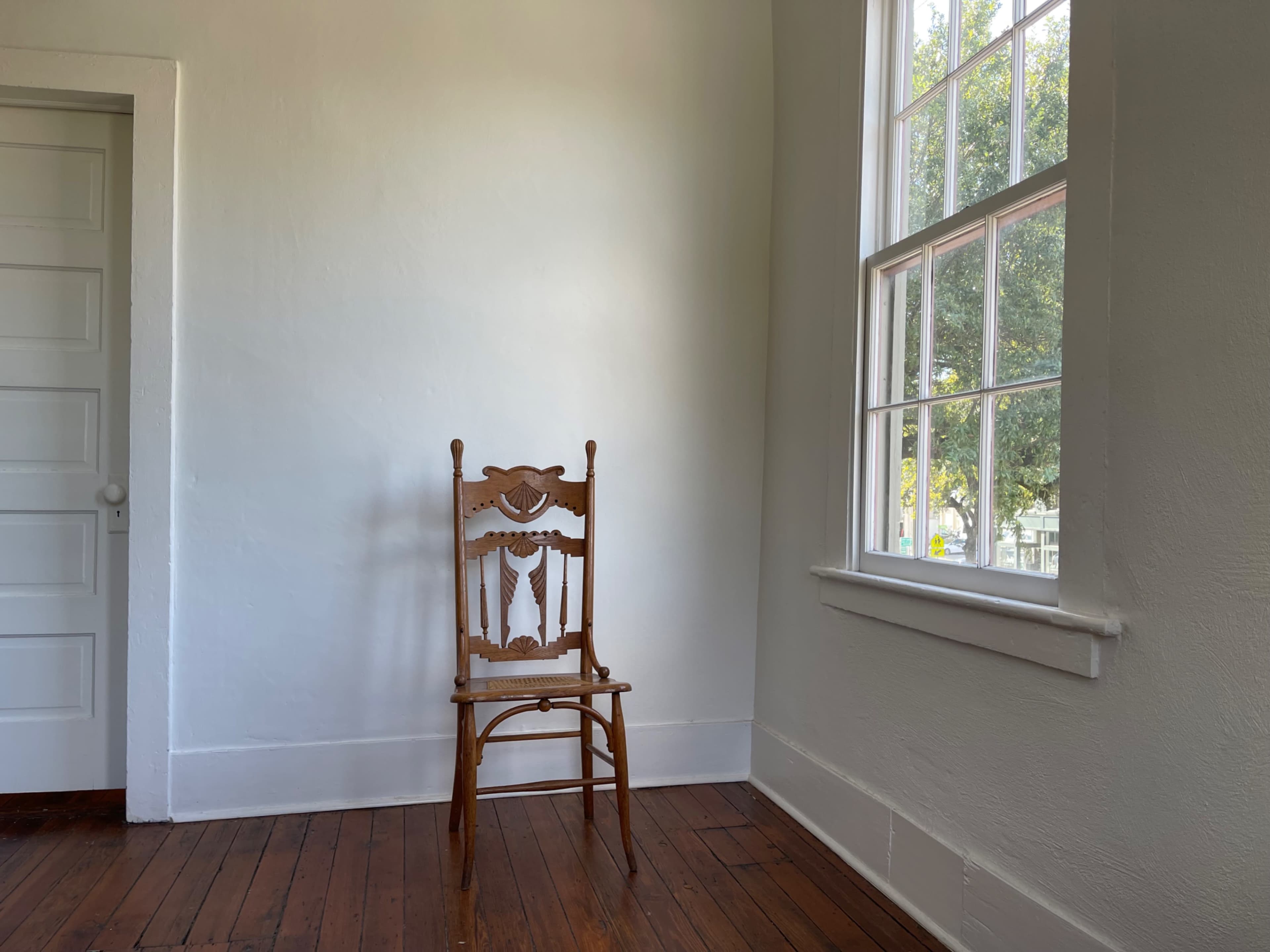A wooden chair is positioned against a white wall in a room with a window and a door.
