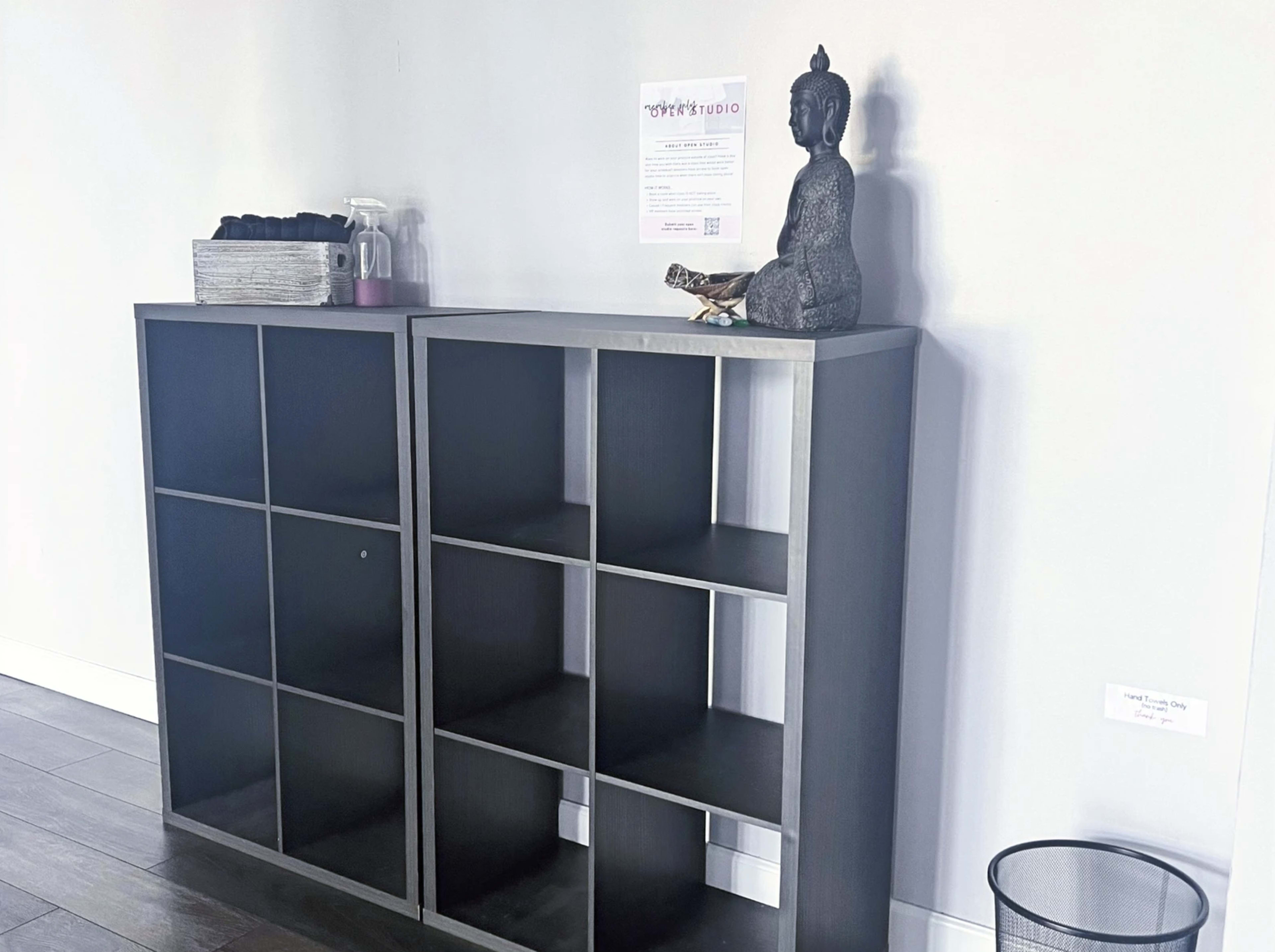Two gray shelving units with open compartments, a decorative Buddha statue on top, and a small trash can beside them.