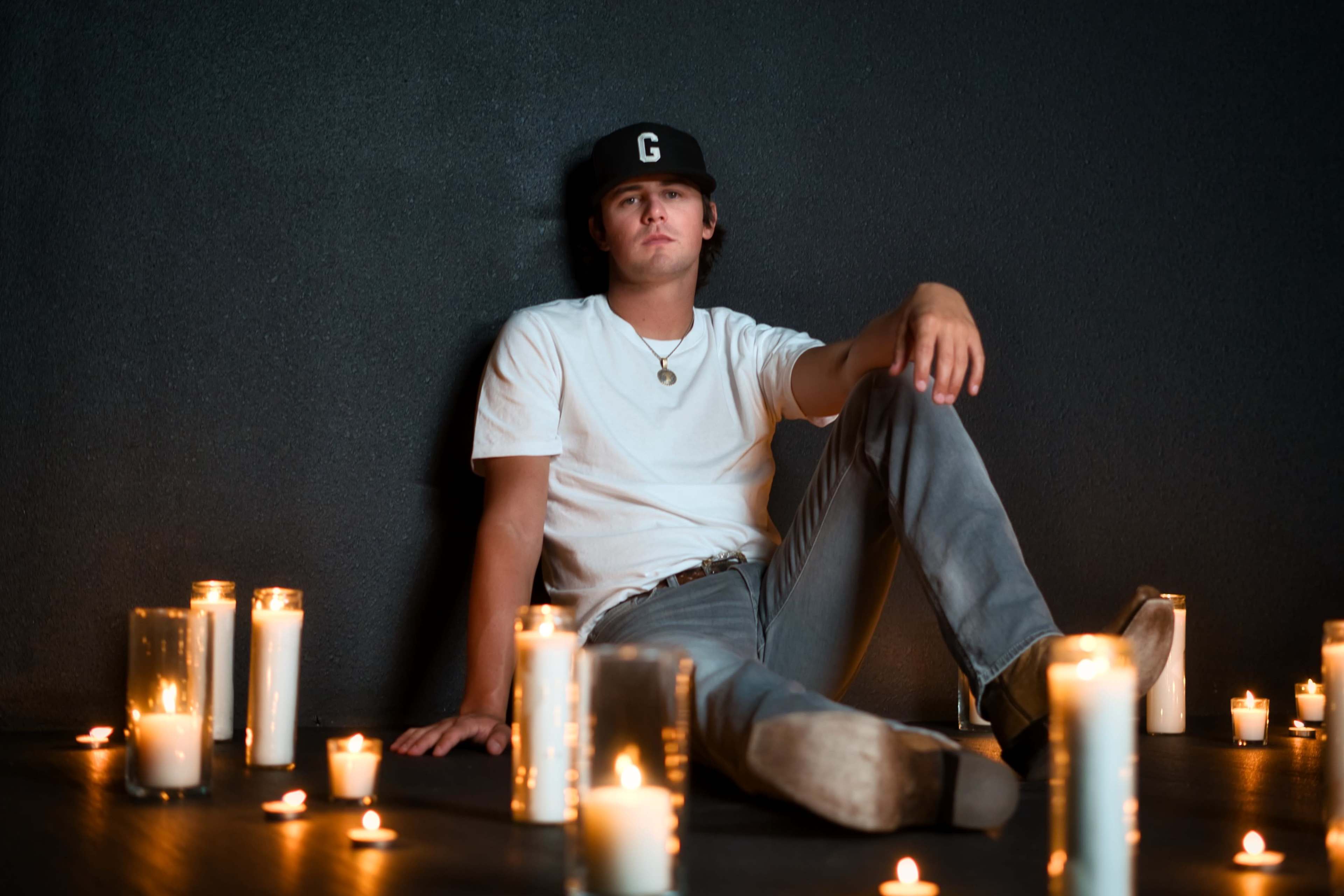 A person wearing a white shirt and a black cap sits on the floor surrounded by lit candles.
