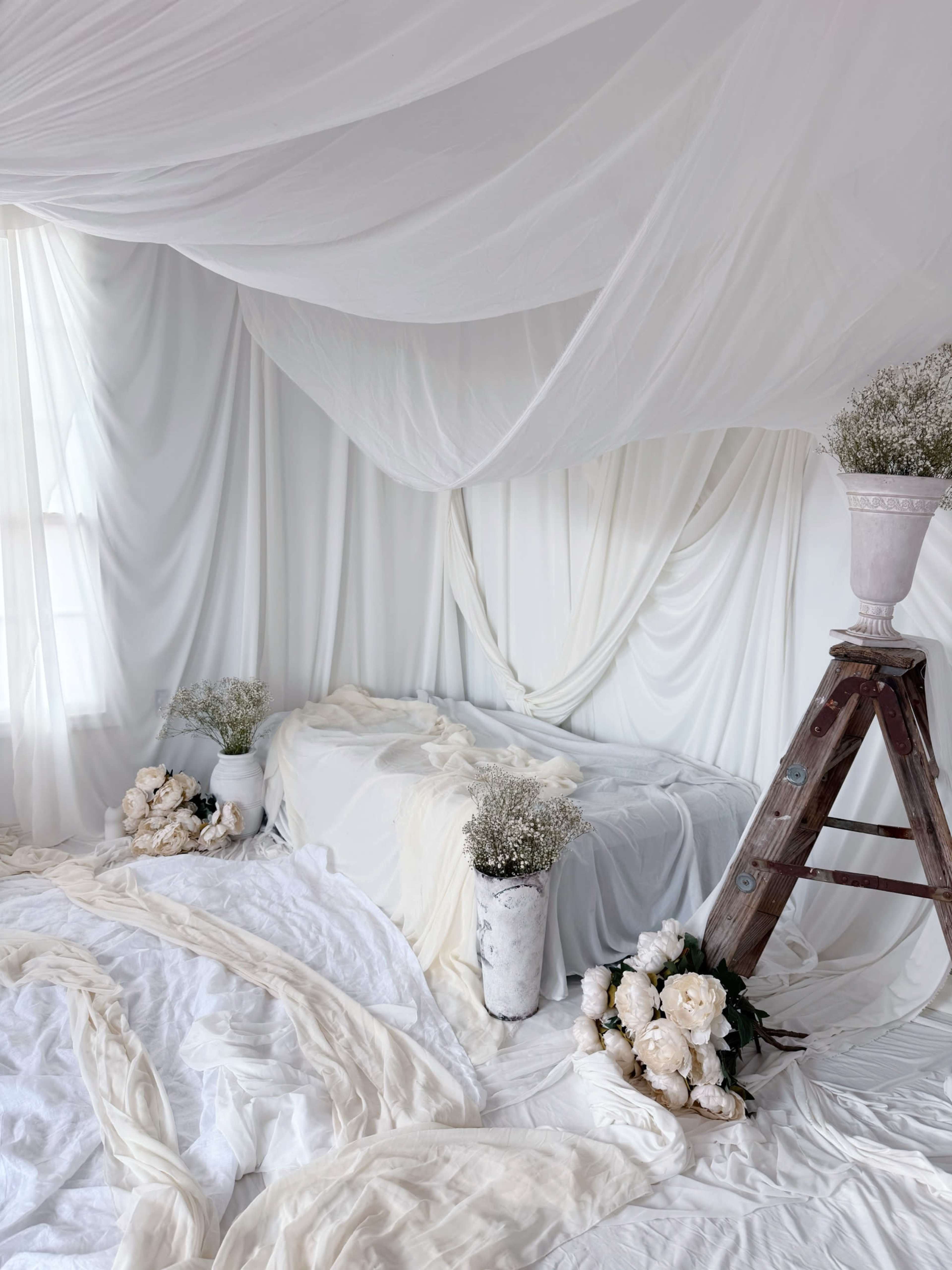A softly draped white room with a bed covered in flowing fabrics, adorned with flowers in vases and positioned near a wooden ladder.