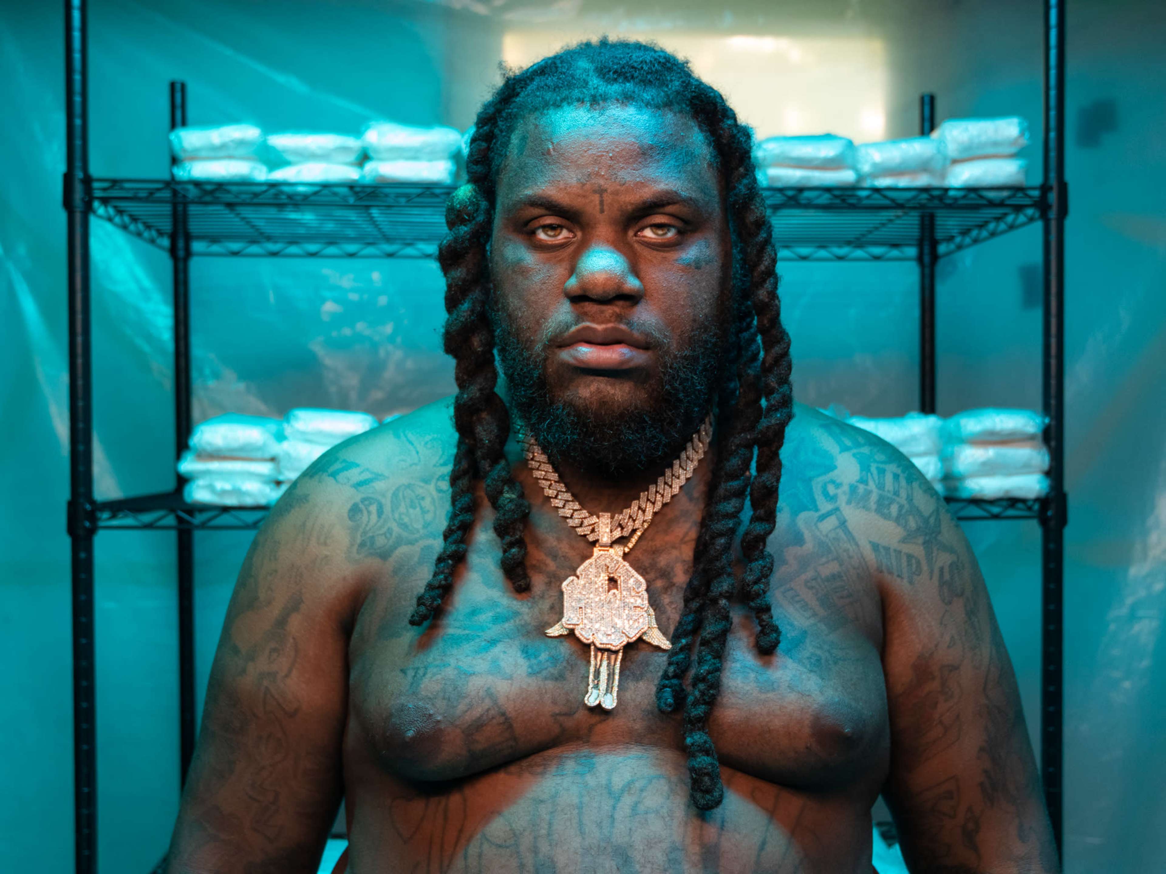 A shirtless man with long braids stands in front of stacked bags on shelves within a brightly lit room.