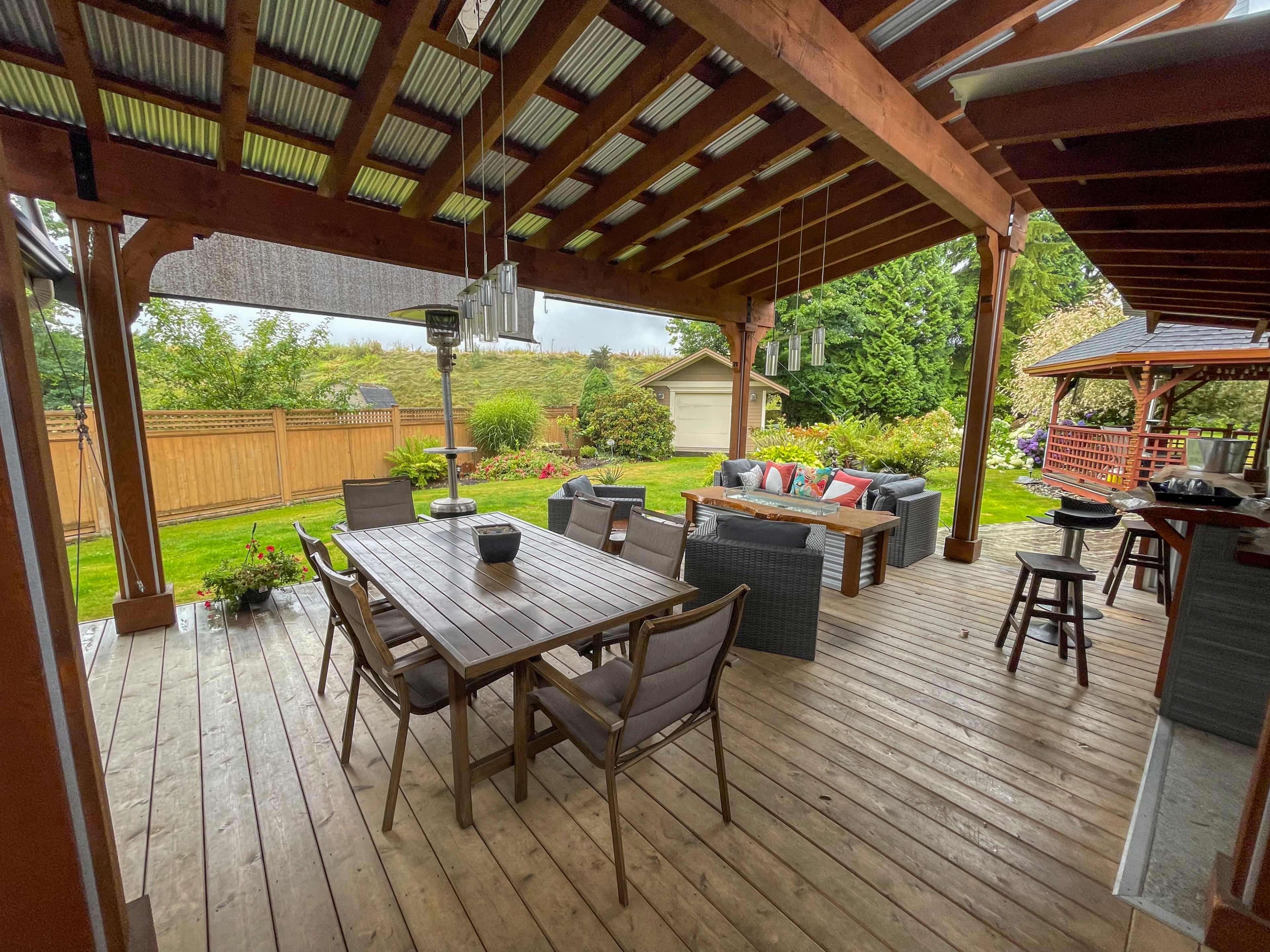 A covered outdoor patio area with a wooden dining table, chairs, and a seating arrangement, surrounded by greenery and a shed in the background.