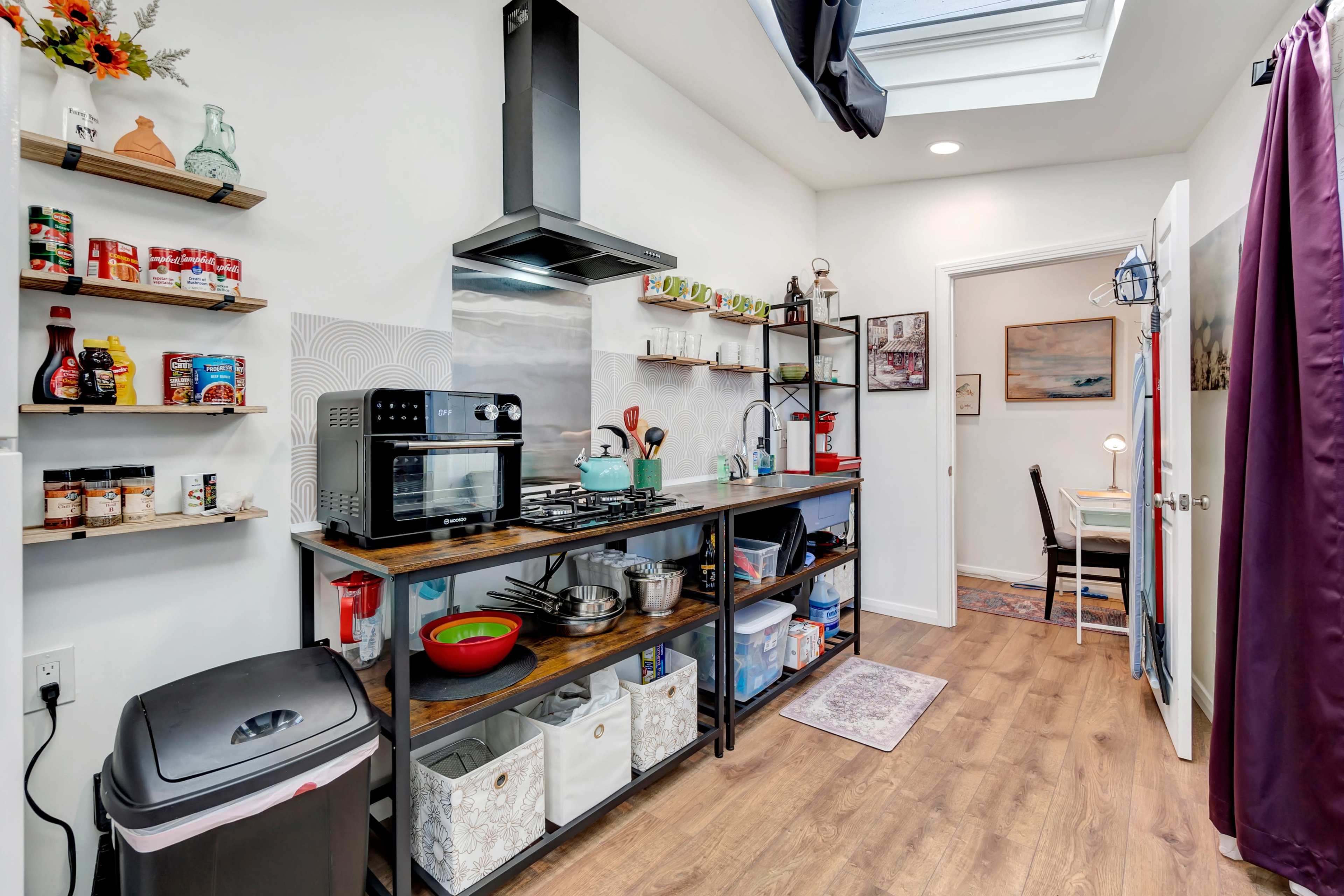 The image shows a modern kitchen with an oven, gas stove, open shelving, and organized storage bins, complemented by a skylight and a dining area visible through a doorway.