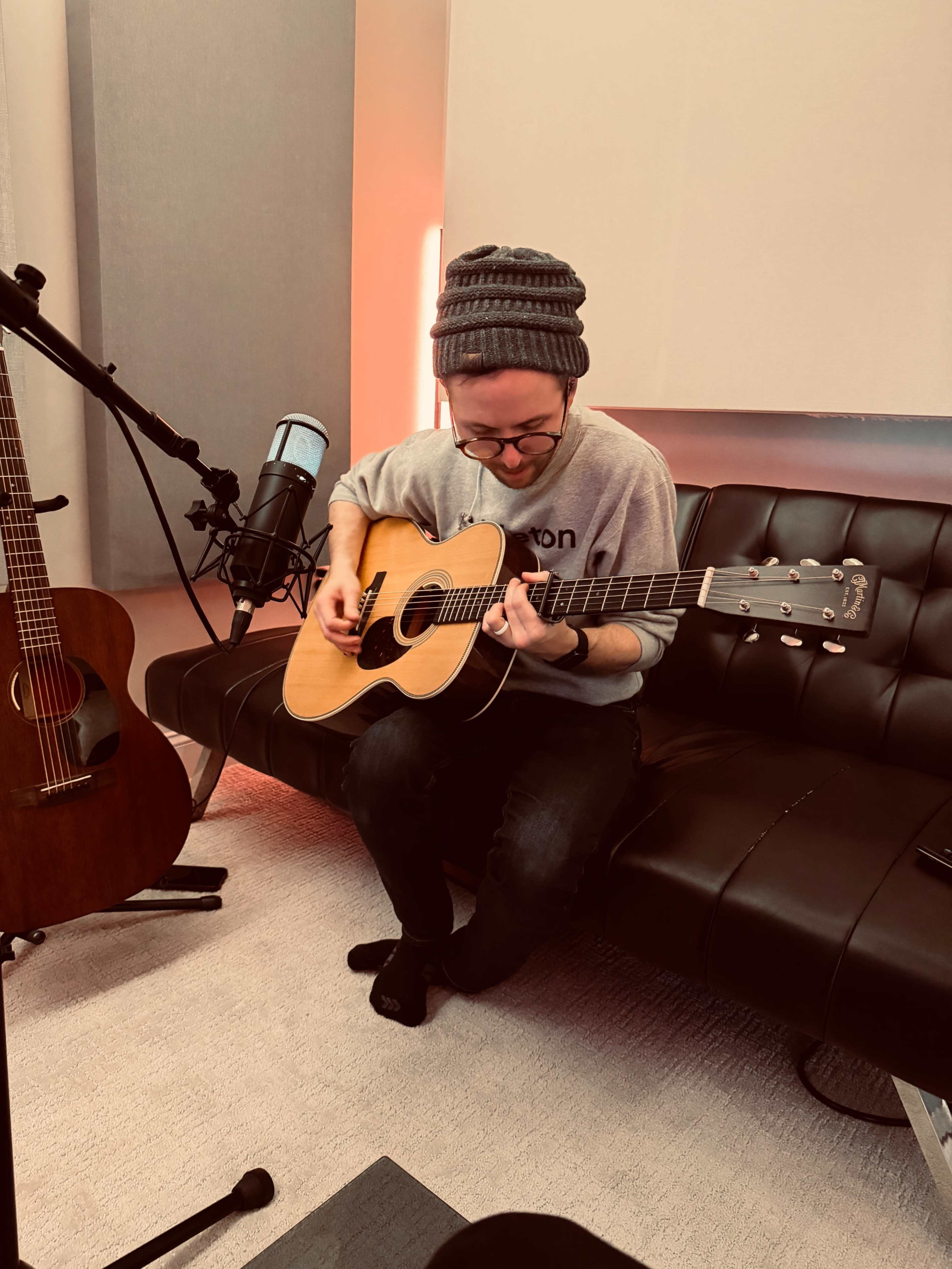 A person wearing glasses and a beanie plays an acoustic guitar while seated on a black sofa in a studio setting.