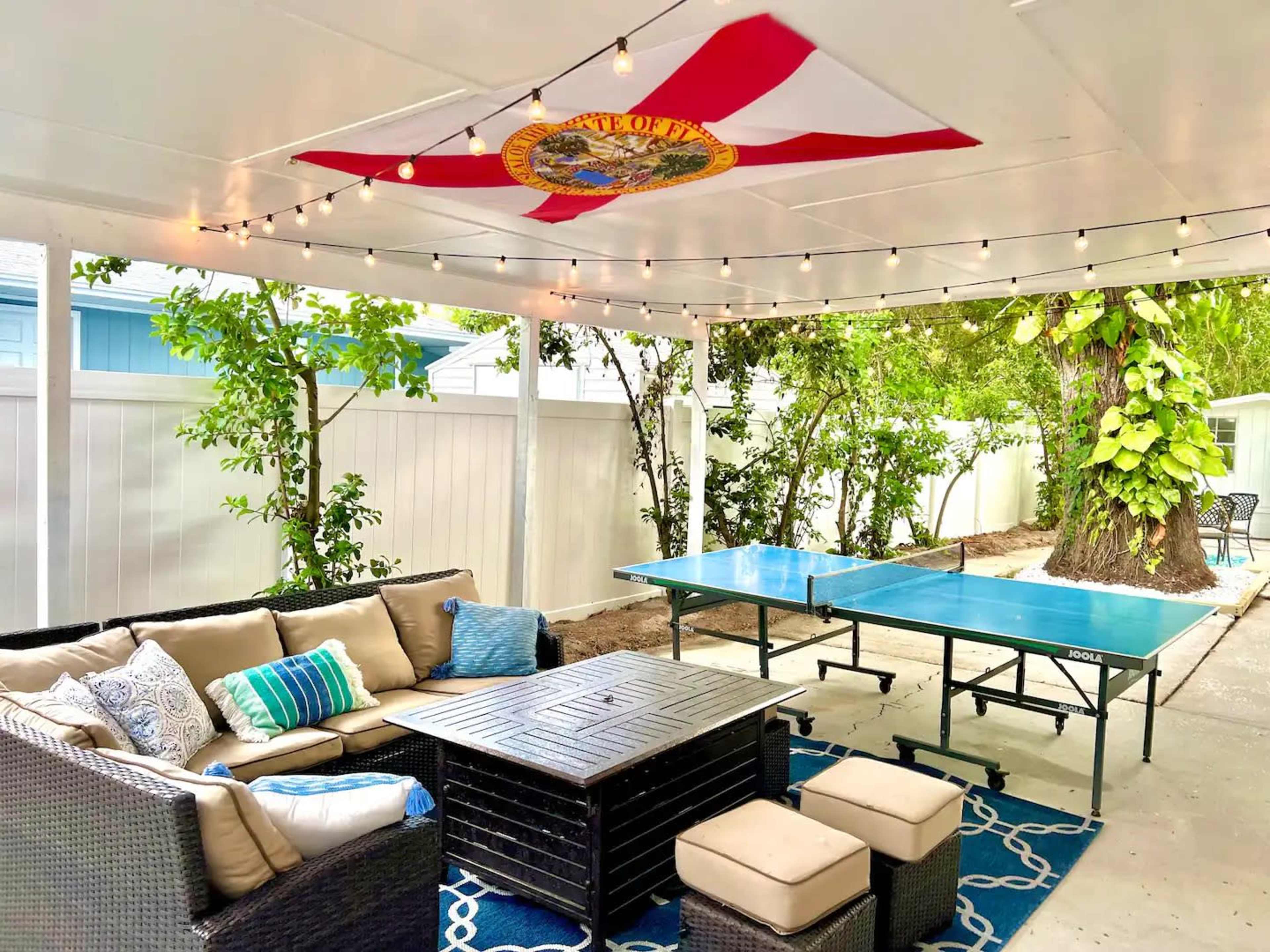 A covered outdoor patio features a seating area with a sectional sofa and coffee table, alongside a ping pong table under string lights and a flag hanging from the ceiling.