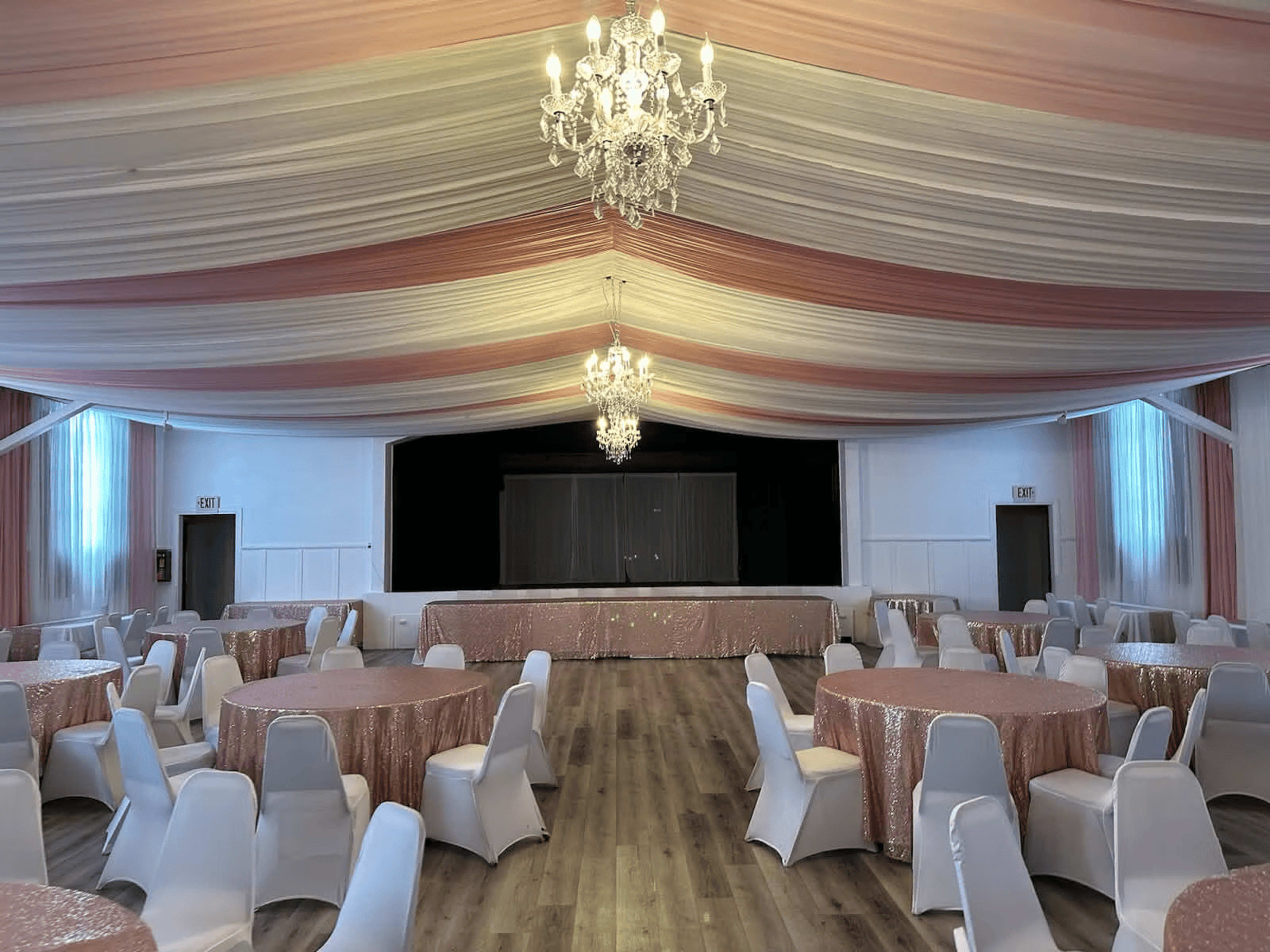 The interior of a banquet hall features round tables with white chairs and a staged area at the front, all accented by draped fabric and chandeliers overhead.