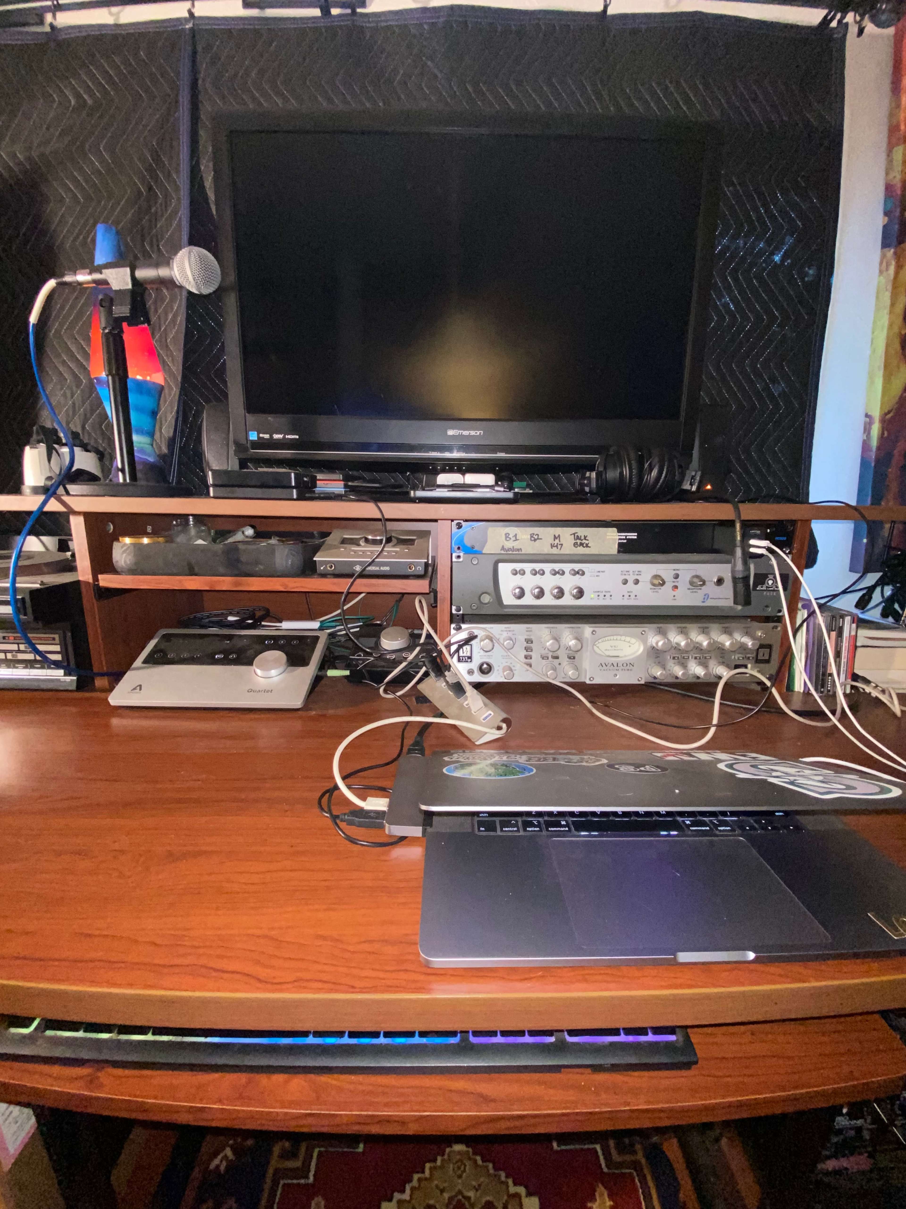 A wooden desk is set up with a laptop, audio equipment, a microphone, and a television, all surrounded by various cables and accessories.