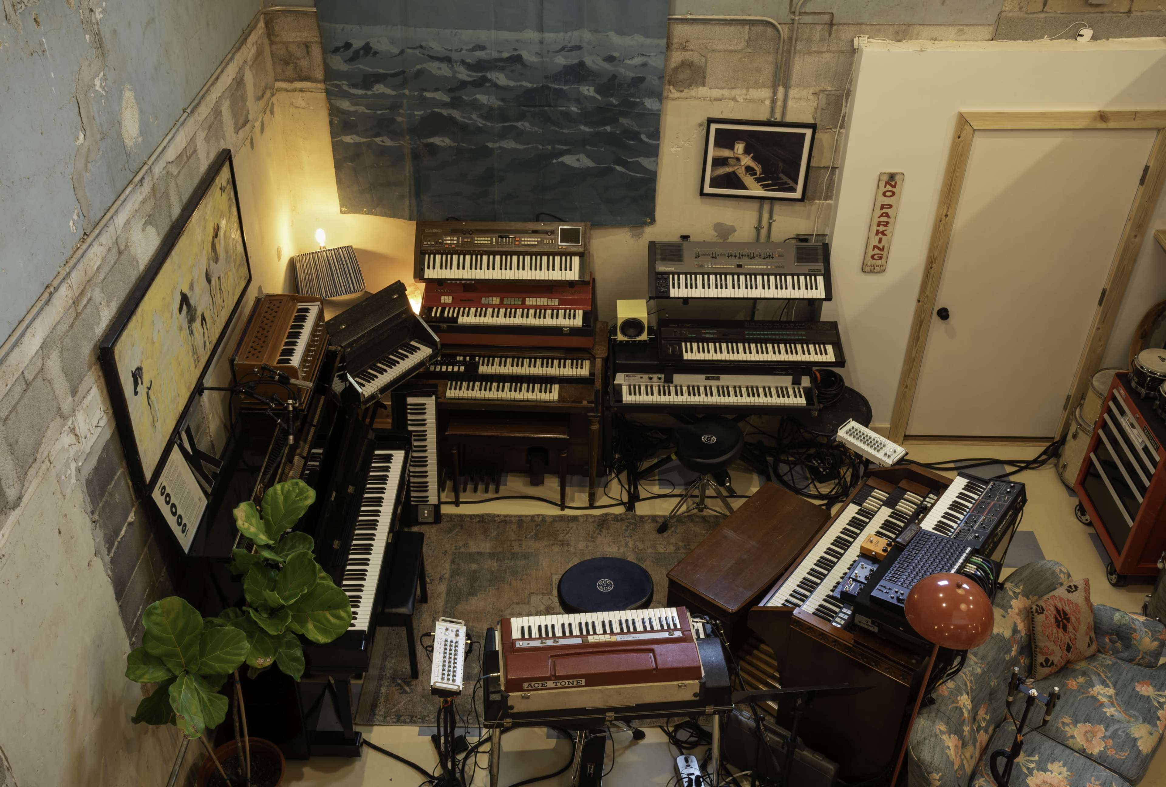 The image shows a music studio filled with various keyboards and synthesizers arranged in a cozy, cluttered space, with a large blue wall hanging and a green plant in the corner.