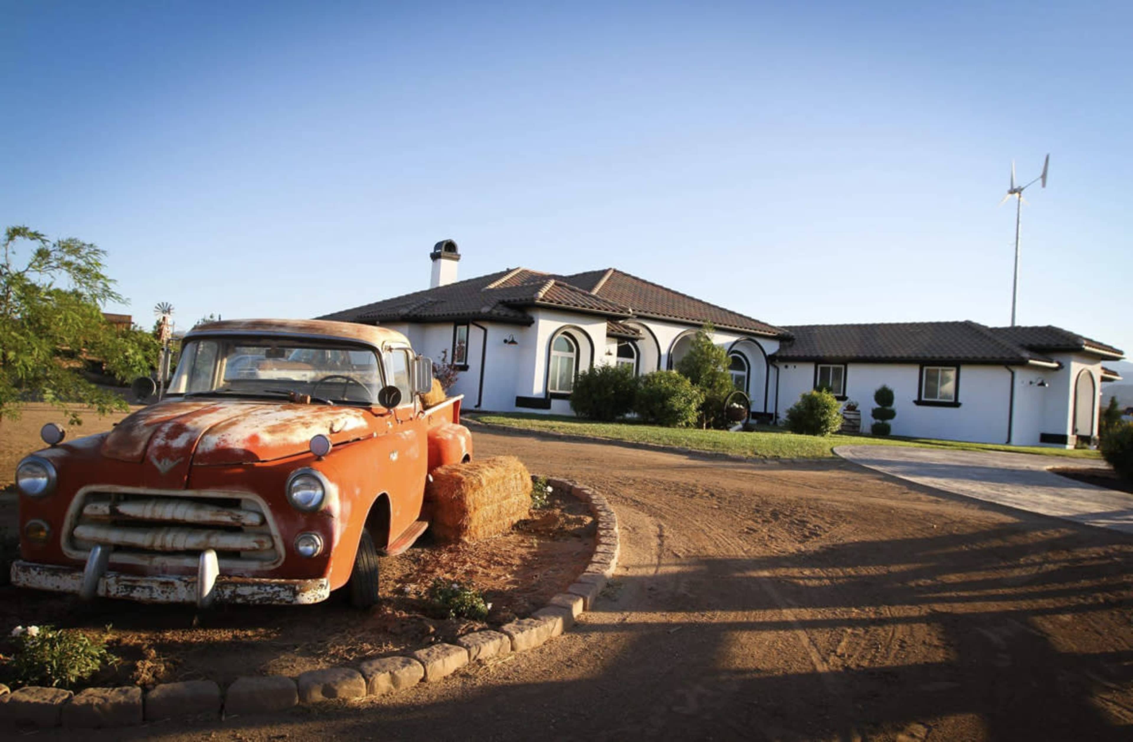An old orange truck is parked beside a winding driveway leading to a modern white house with a tiled roof.