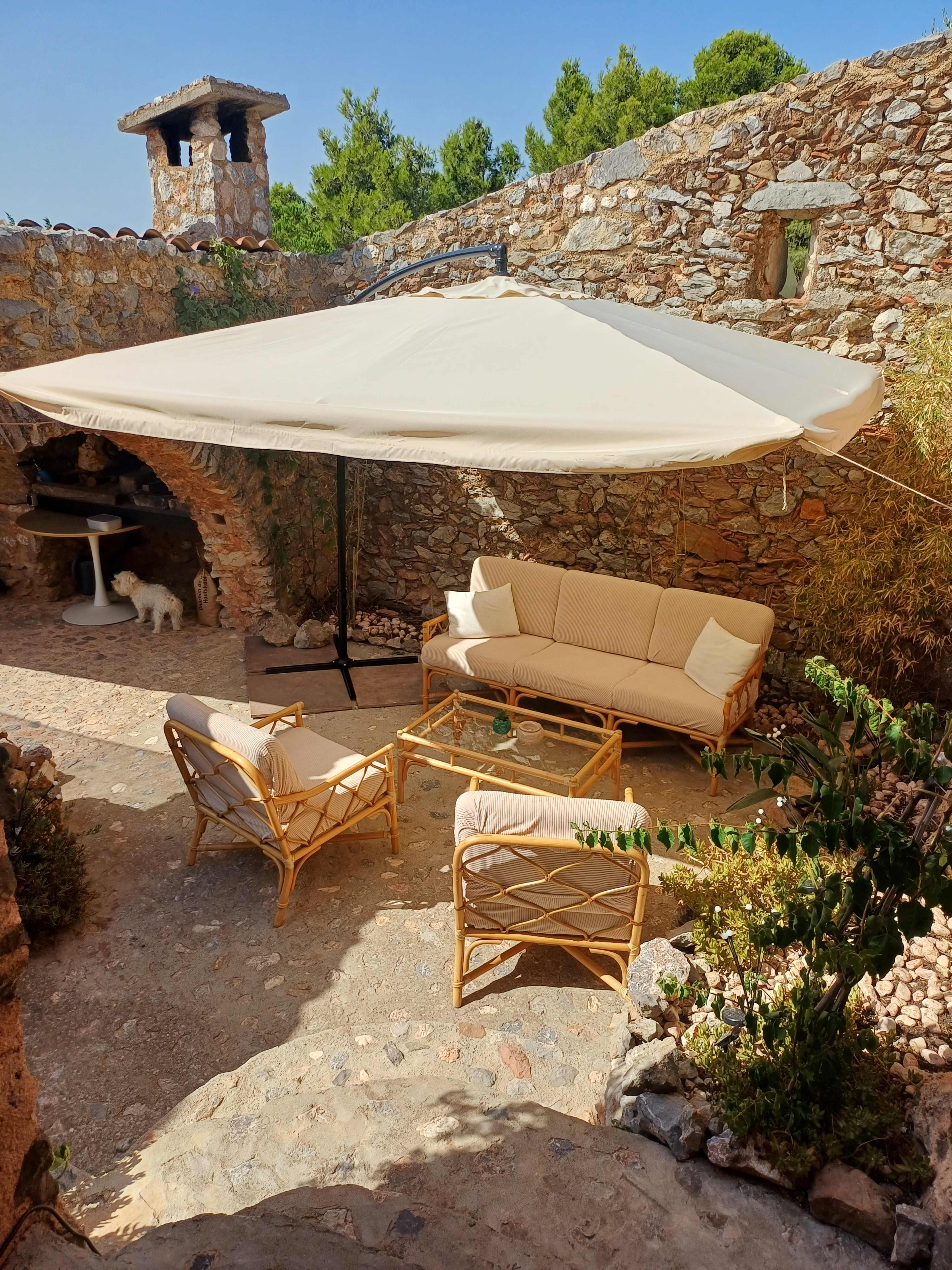 The image shows a cozy outdoor seating area with a large umbrella, surrounded by stone walls, featuring a rattan sofa, chairs, and a coffee table.