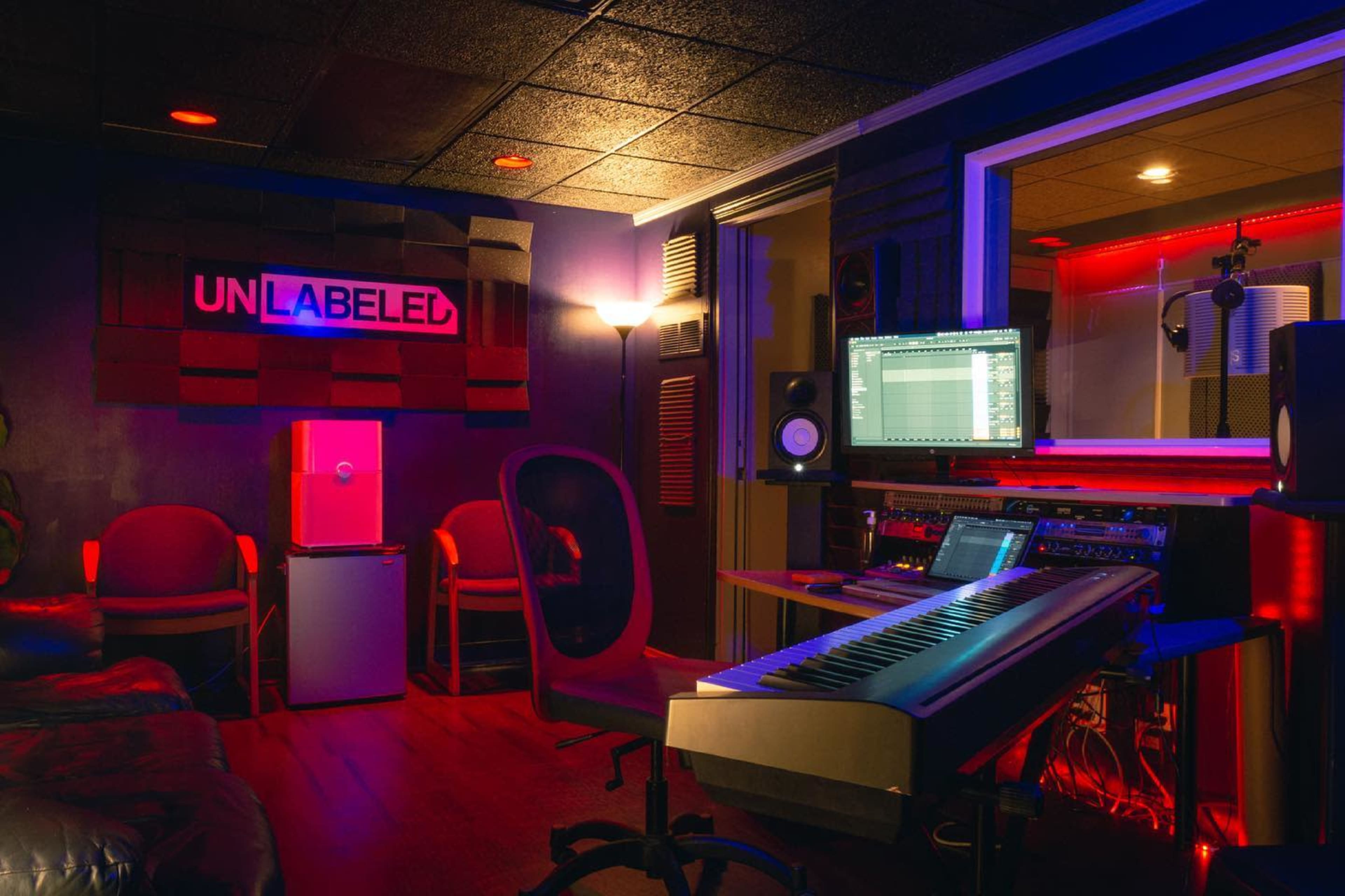 A music studio features a keyboard, computer setup, and red-lit decor, with soundproofing panels on the walls and two chairs in the corner.