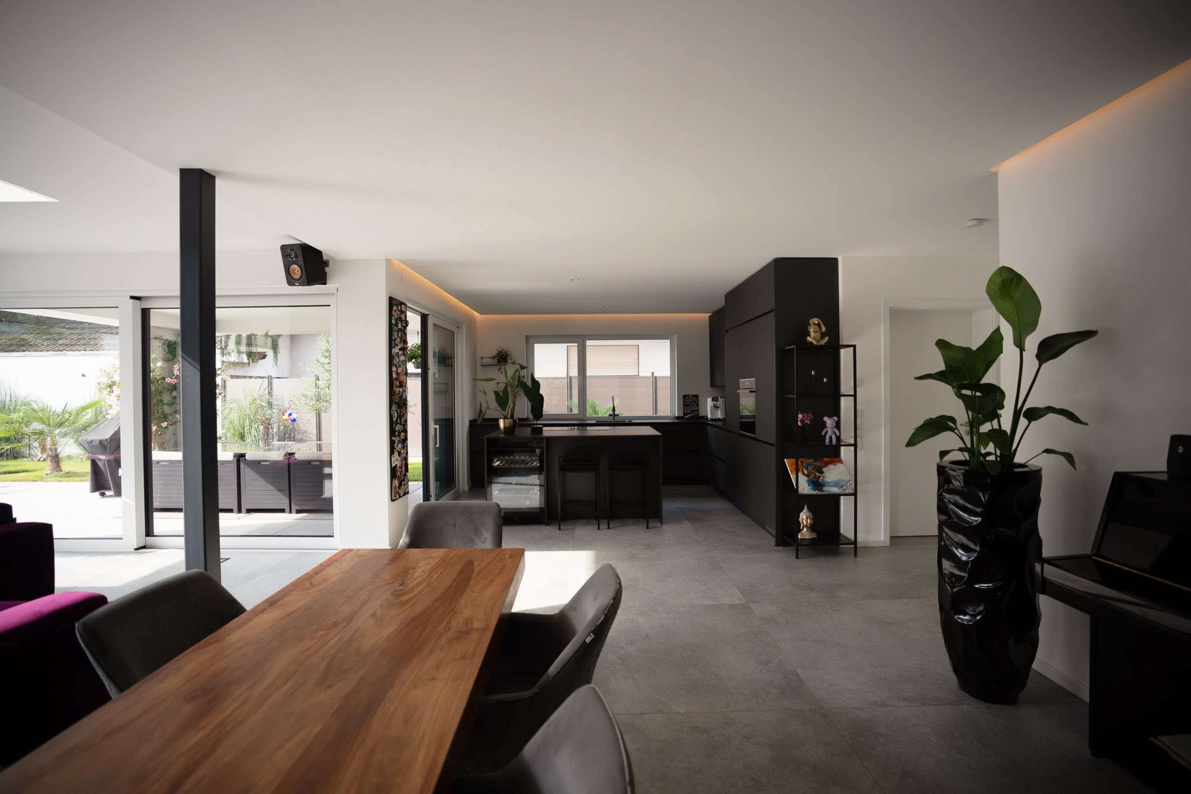 The image shows a modern living space with a dining area featuring a wooden table, black chairs, and an open kitchen in the background, complemented by large windows that lead to an outdoor area.